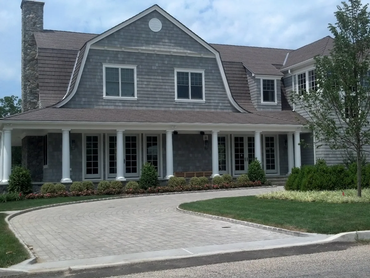 Large gray shingle-style home with a gambrel roof in an Enviroshake composite shake, columned front porch, stone chimney, and a circular paved driveway.