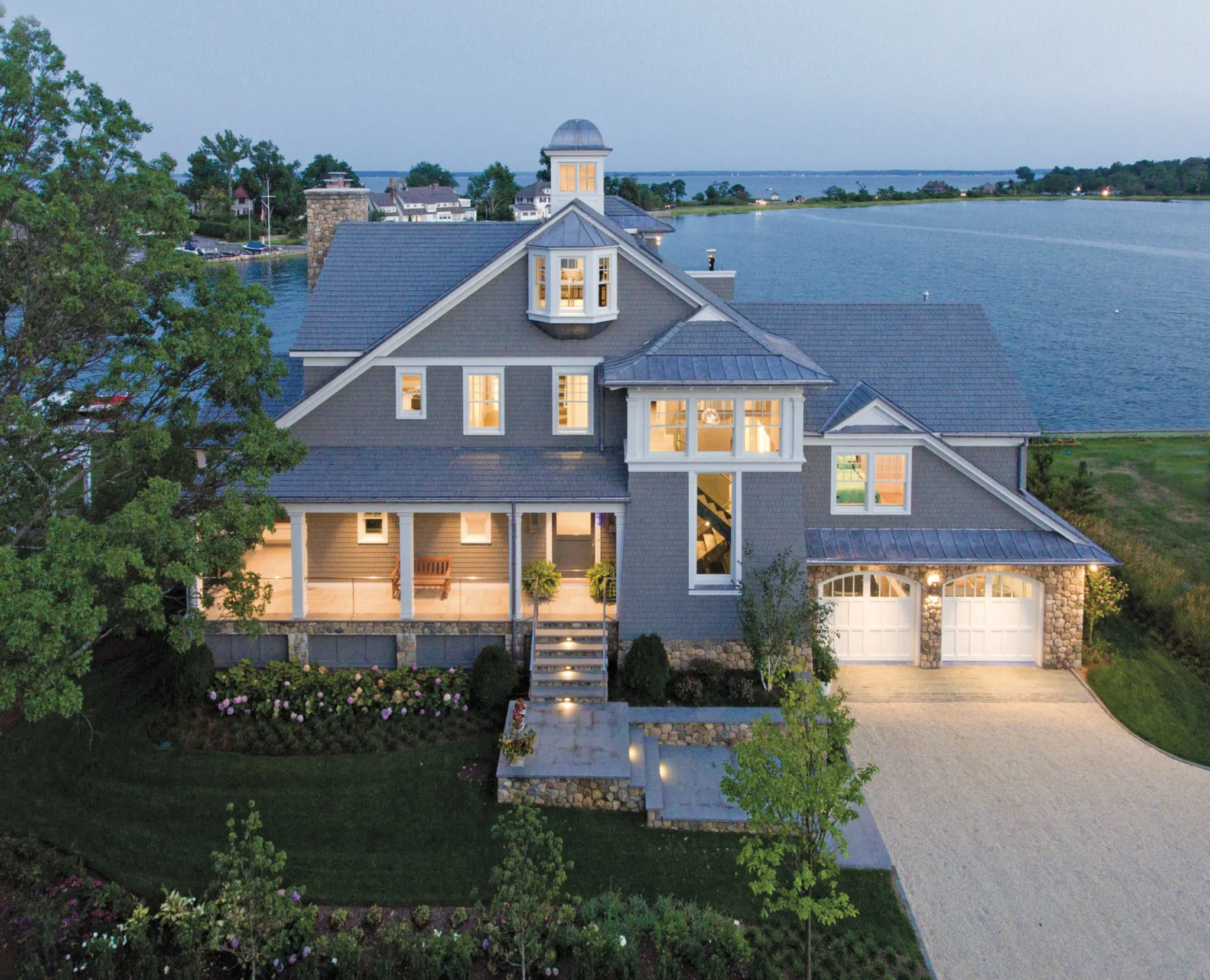 Twilight aerial view of a large gray shingle-style waterfront home with an Enviroshake composite roof, cupola, stone accents, and illuminated windows, situated along a coastal inlet.