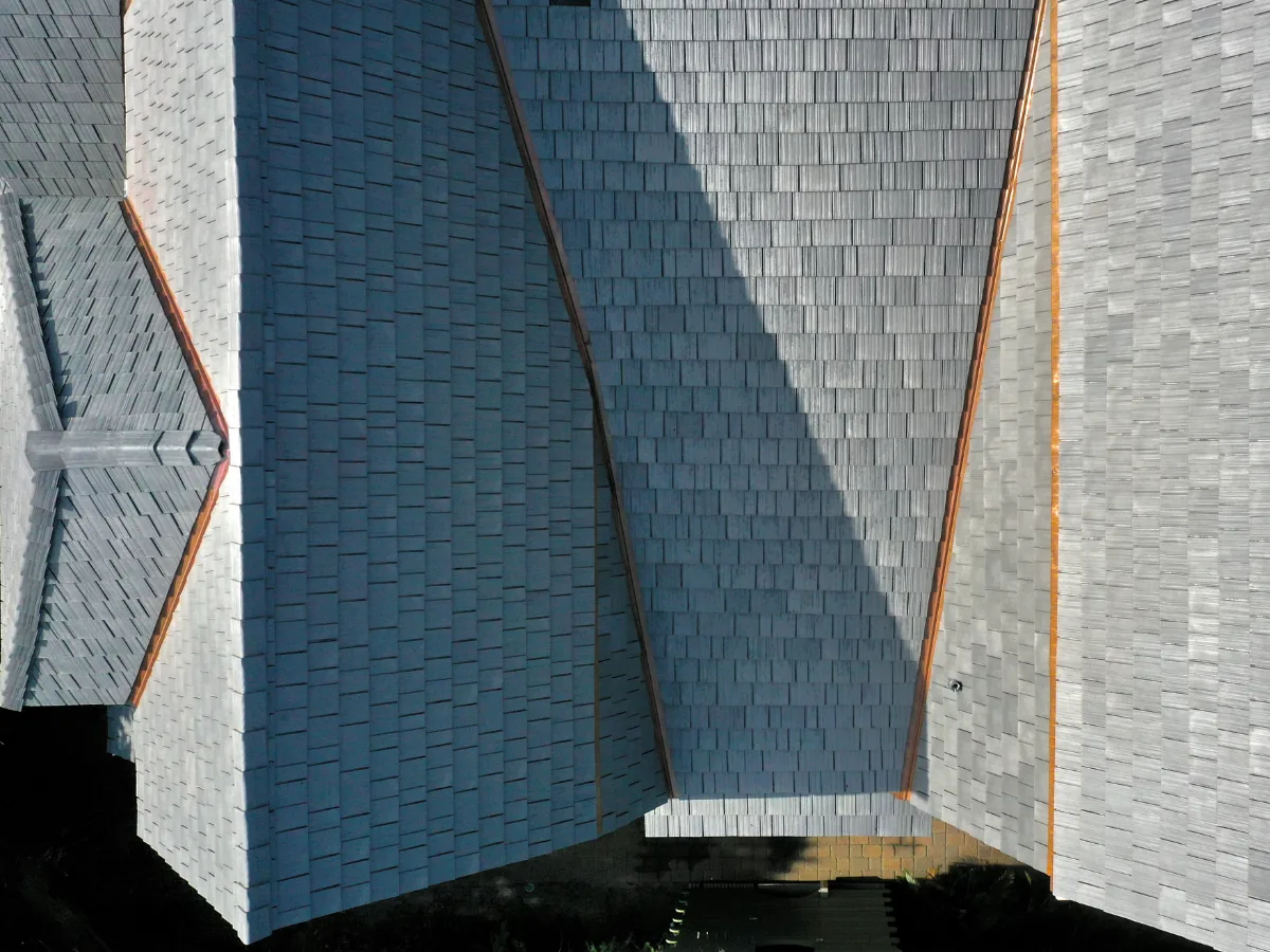 Aerial close-up of multiple gabled and hipped roofs in silvered cedar Enviroshake composite shake, with copper valley flashing and strong sunlight casting sharp shadows.