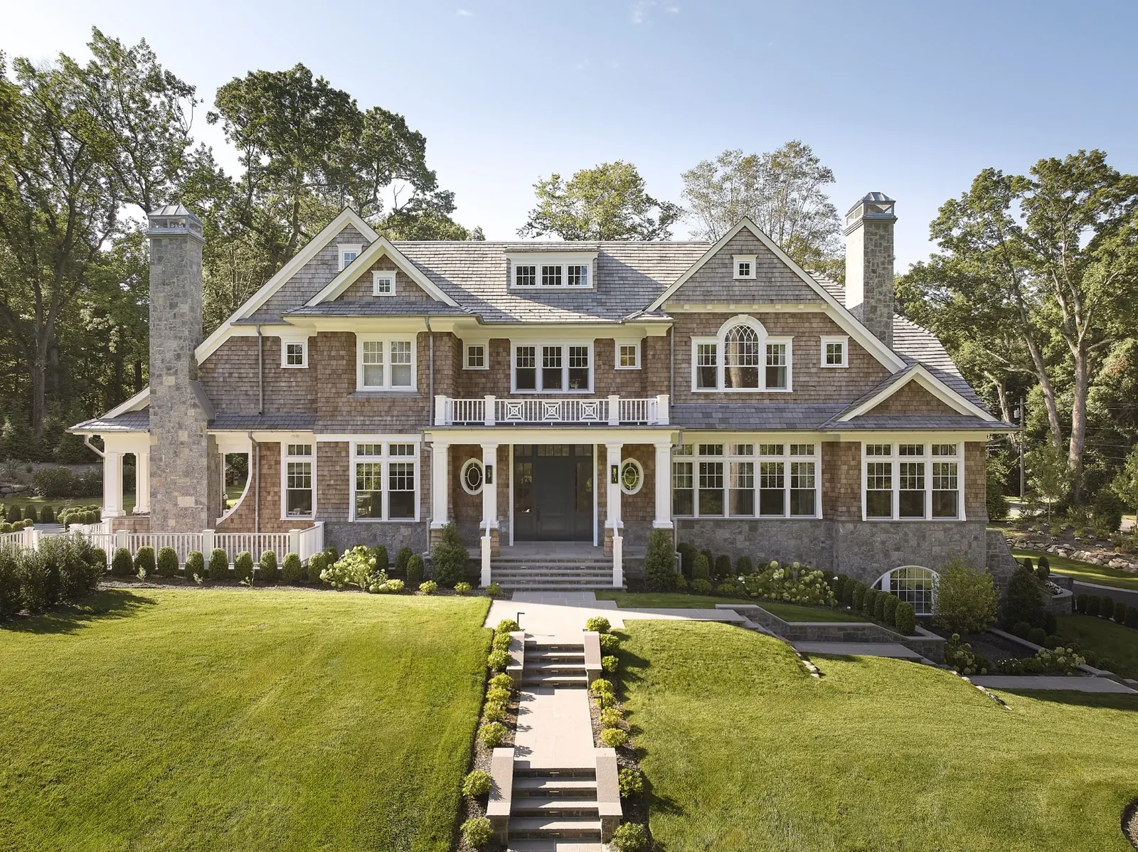 Large shingle-style home with a gray Enviroshake composite roof, cedar shake siding, stone chimneys, and a grand front lawn with landscaped pathway.