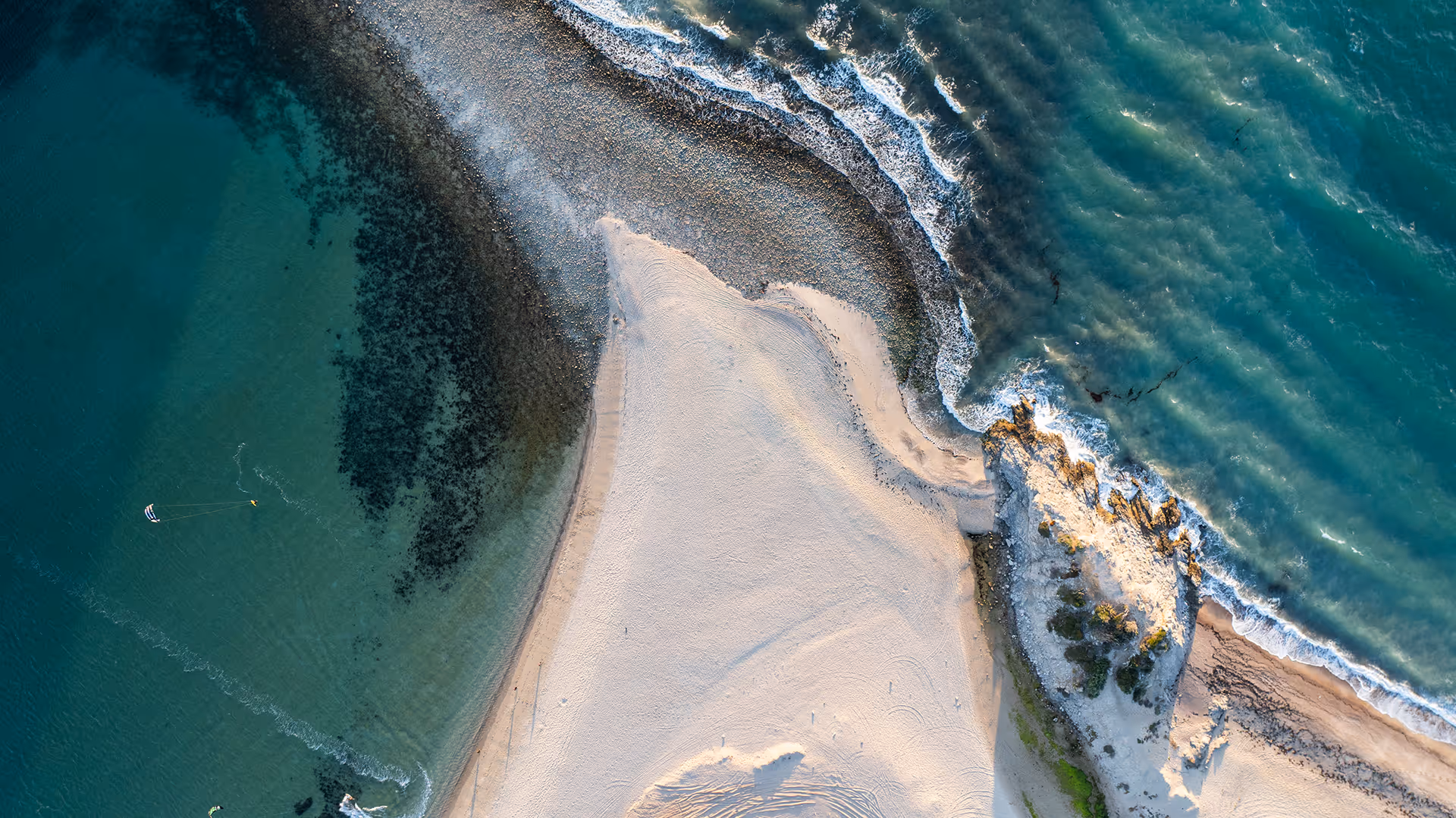 Aerial view of a sandy beach meeting rocky shorelines with waves crashing in clear blue-green ocean water.