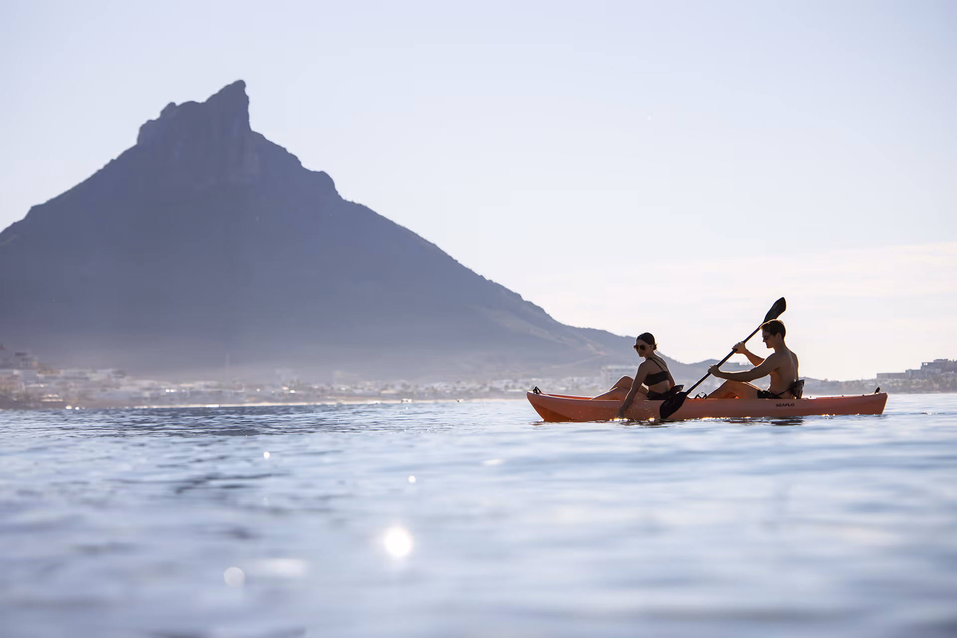 Dos personas remando en kayak naranja sobre agua tranquila con montaña al fondo.