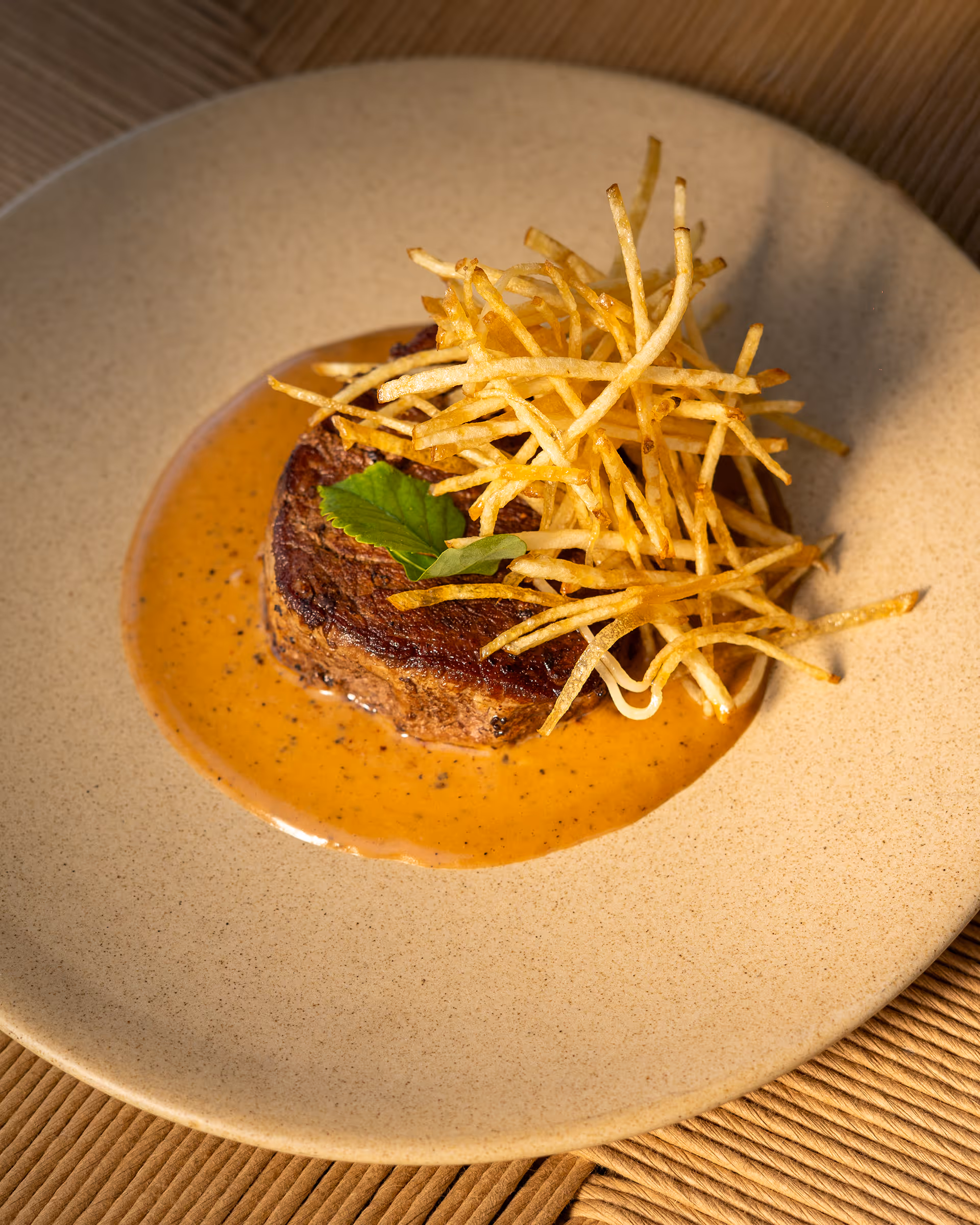 Filete de carne en salsa marrón con un toque de hojas verdes y crujientes papas hilo encima, servido en plato beige.