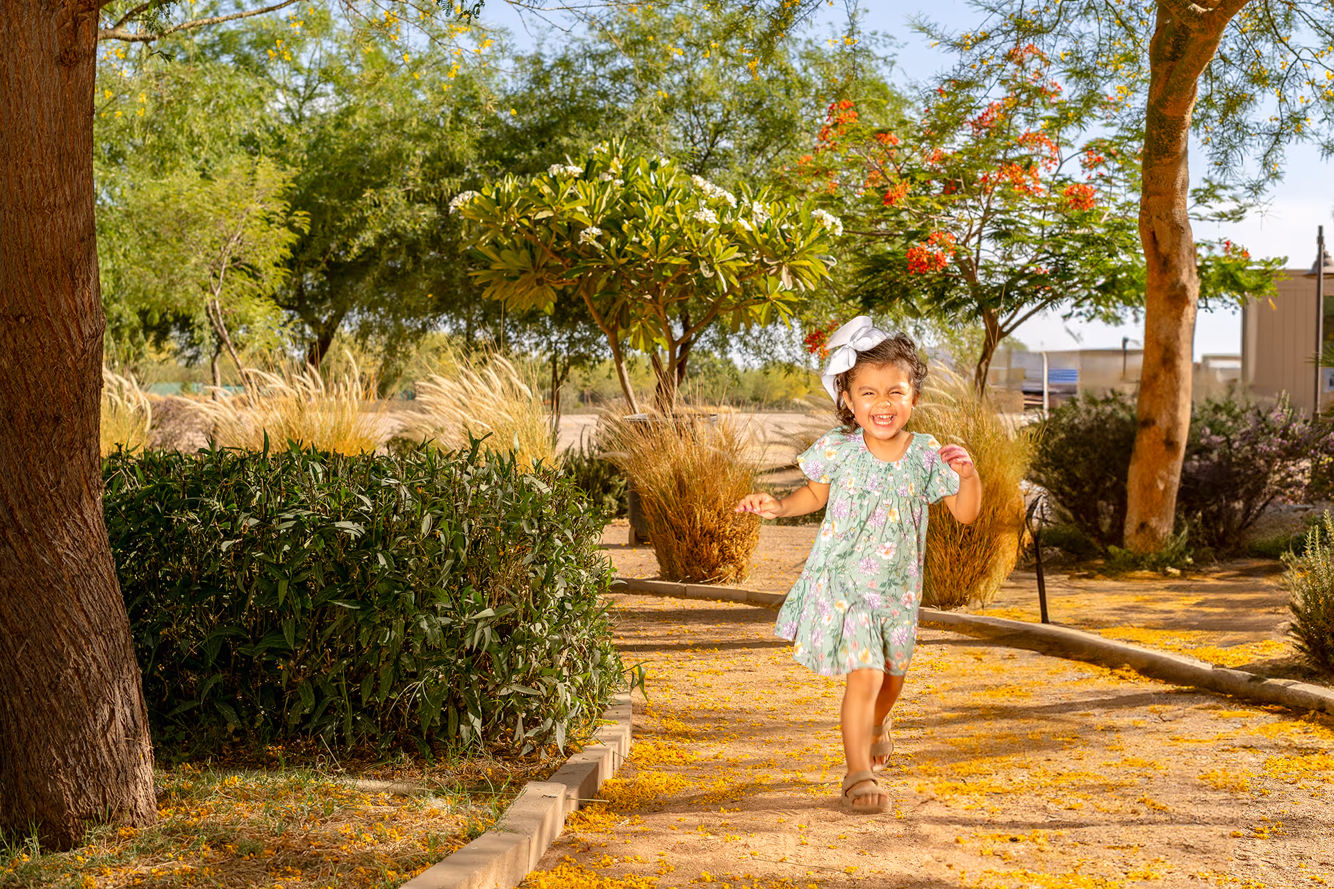 Niña pequeña con vestido floral y moño blanco sonriendo y corriendo por un camino en un parque soleado con árboles y arbustos.