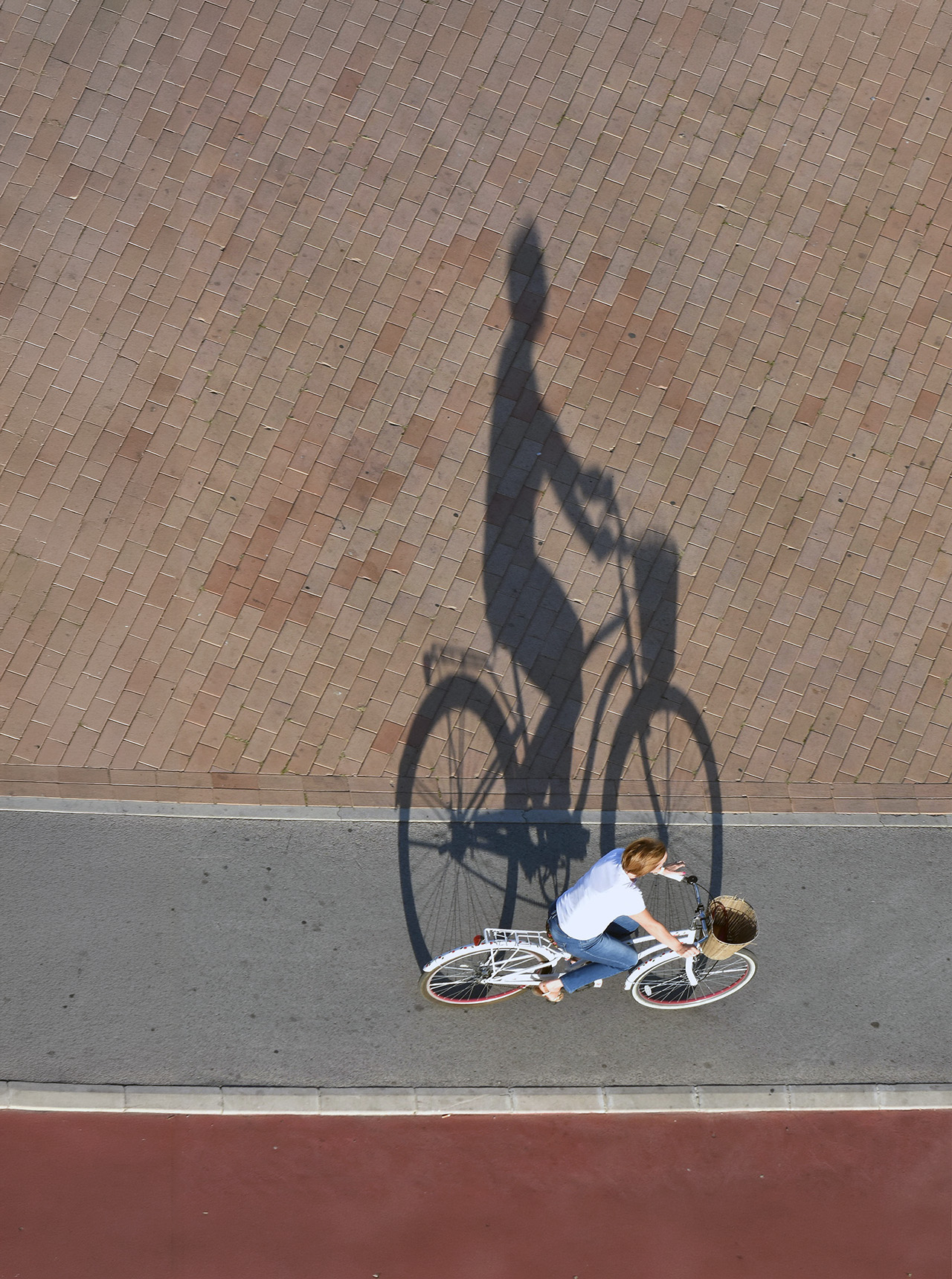 Person riding a white bicycle on a path casting a large shadow on brick pavement.
