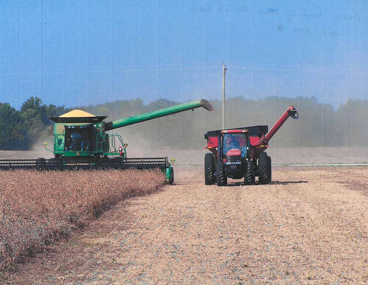Green combine and red tractor harvesting crops in a dusty farm field under a clear sky.