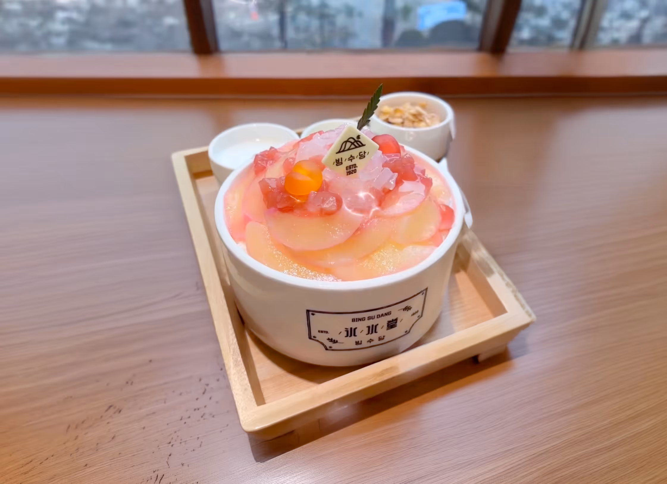 Bingsu dessert topped with peach slices, jelly cubes, and a small leaf, served in a white bowl on a wooden tray with two small side dishes.