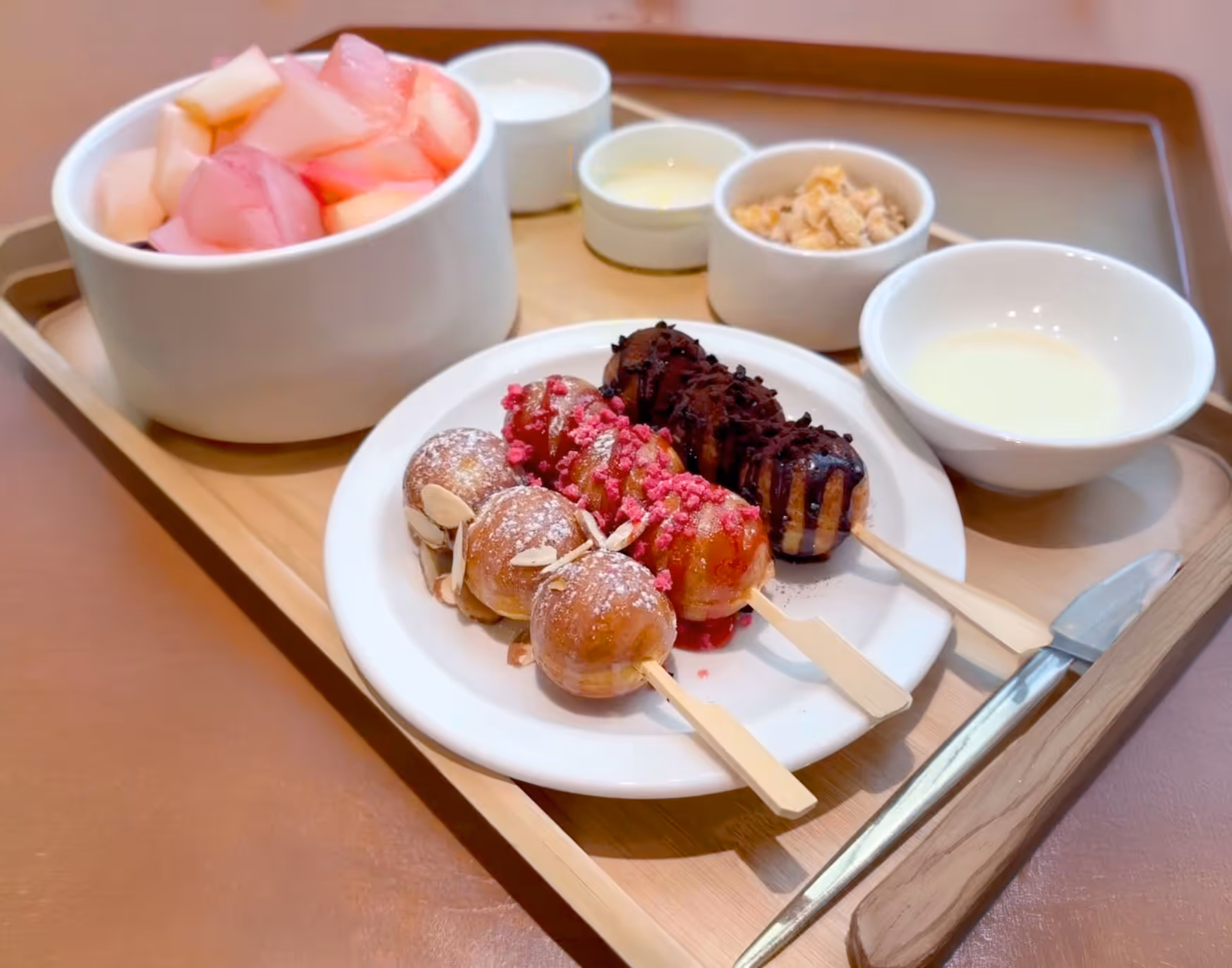 Tray with three skewers of small round pancakes topped with almond slices, pink crumbs, and chocolate, served with bowls of syrup, cream, granola, and fruit pieces.