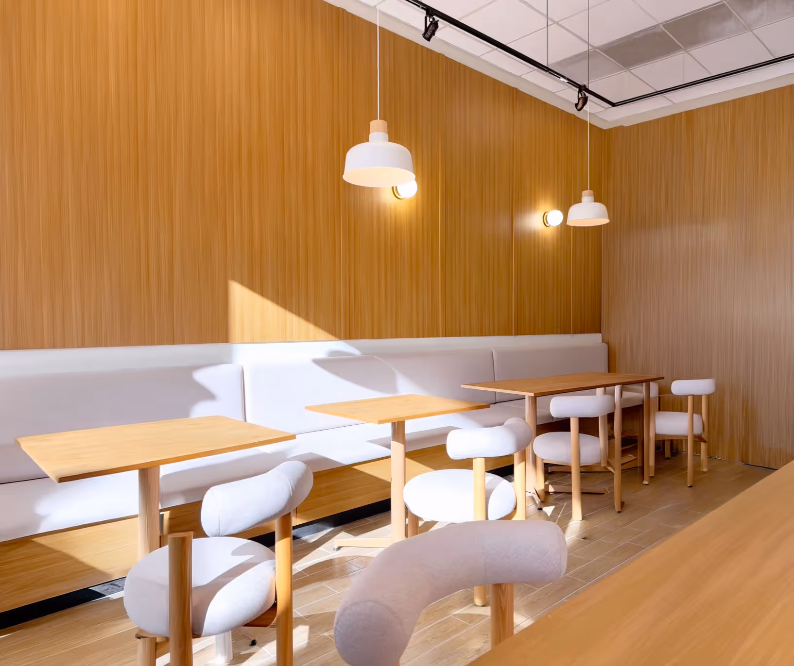 Bright minimalist café interior with wooden tables, light gray cushioned chairs, and white pendant lights against wood-paneled walls.