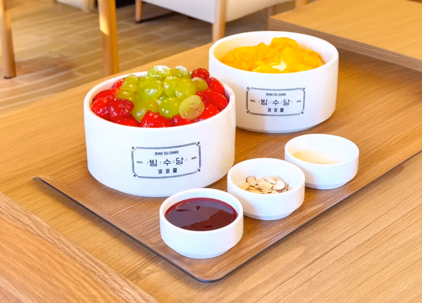 Two bowls of BingsuDang shaved ice desserts with strawberries and grapes in one, mango in the other, served with small side dishes of syrup, sliced almonds, and condensed milk on a wooden tray.