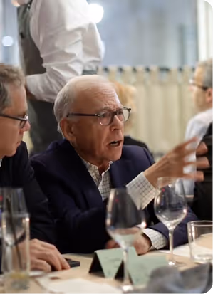 Elderly man with glasses in a suit speaking animatedly at a dining table with others.
