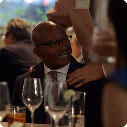 Man in glasses and suit smiling while another person rests a hand on his shoulder at a dinner event.