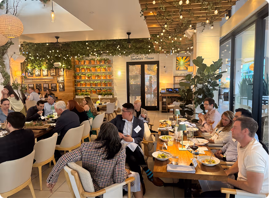 People dining and conversing at a restaurant with hanging lights and green plant decor.