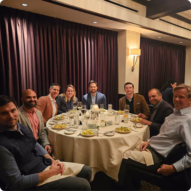 Eight people seated around a round dining table with plates of salad in a dimly lit room.