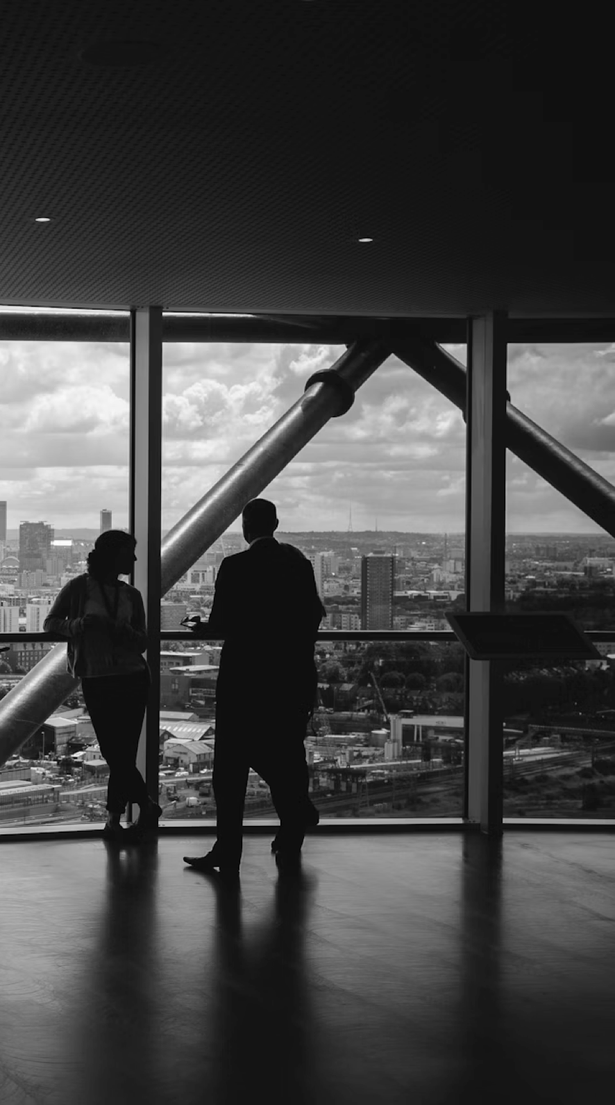 Silhouettes of two people standing by large windows overlooking a cityscape with cloudy sky.