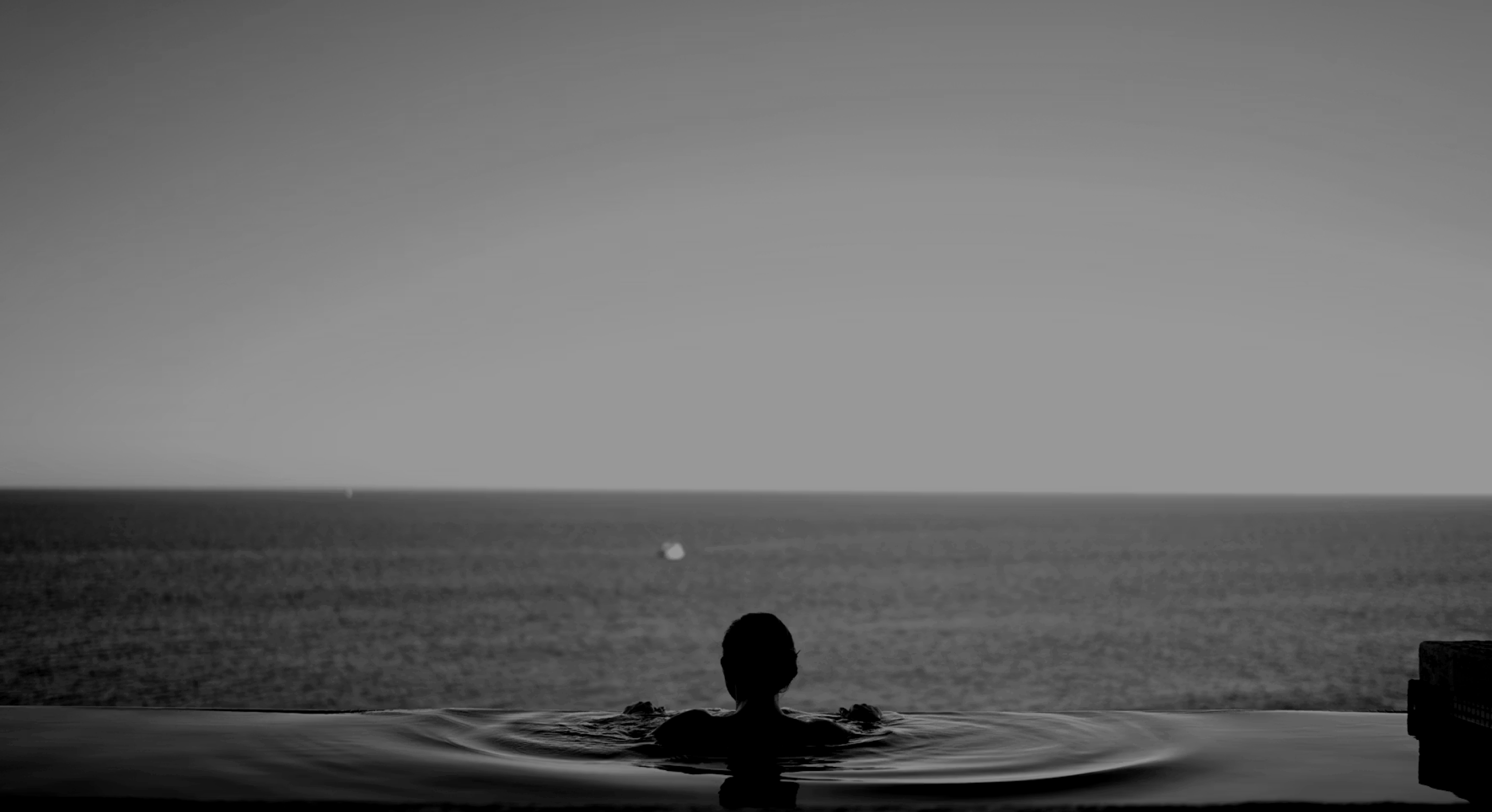 Silhouette of person in pool facing a vast calm ocean under a clear sky.