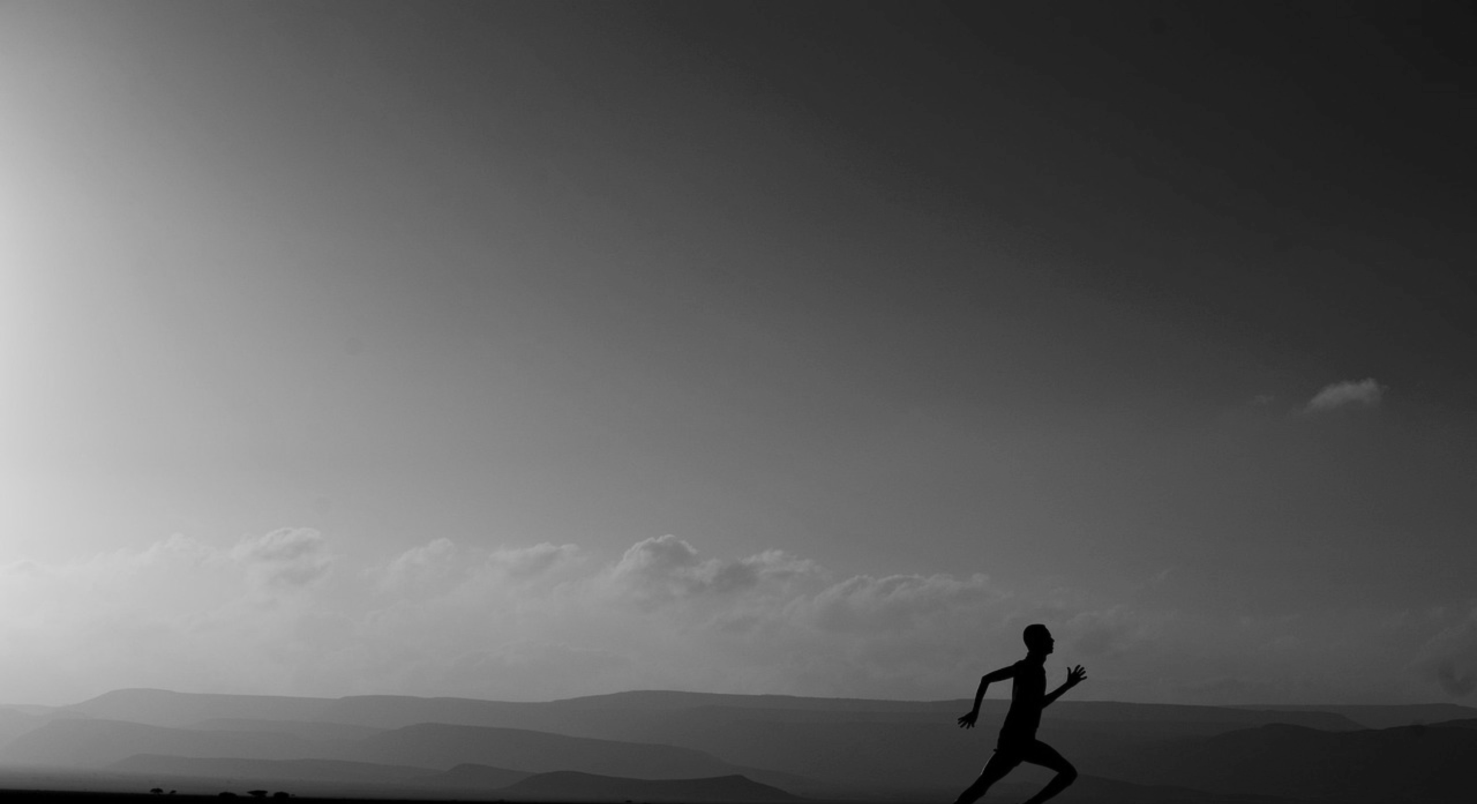 Silhouette of a person running with layered mountain landscape and cloudy sky in the background.