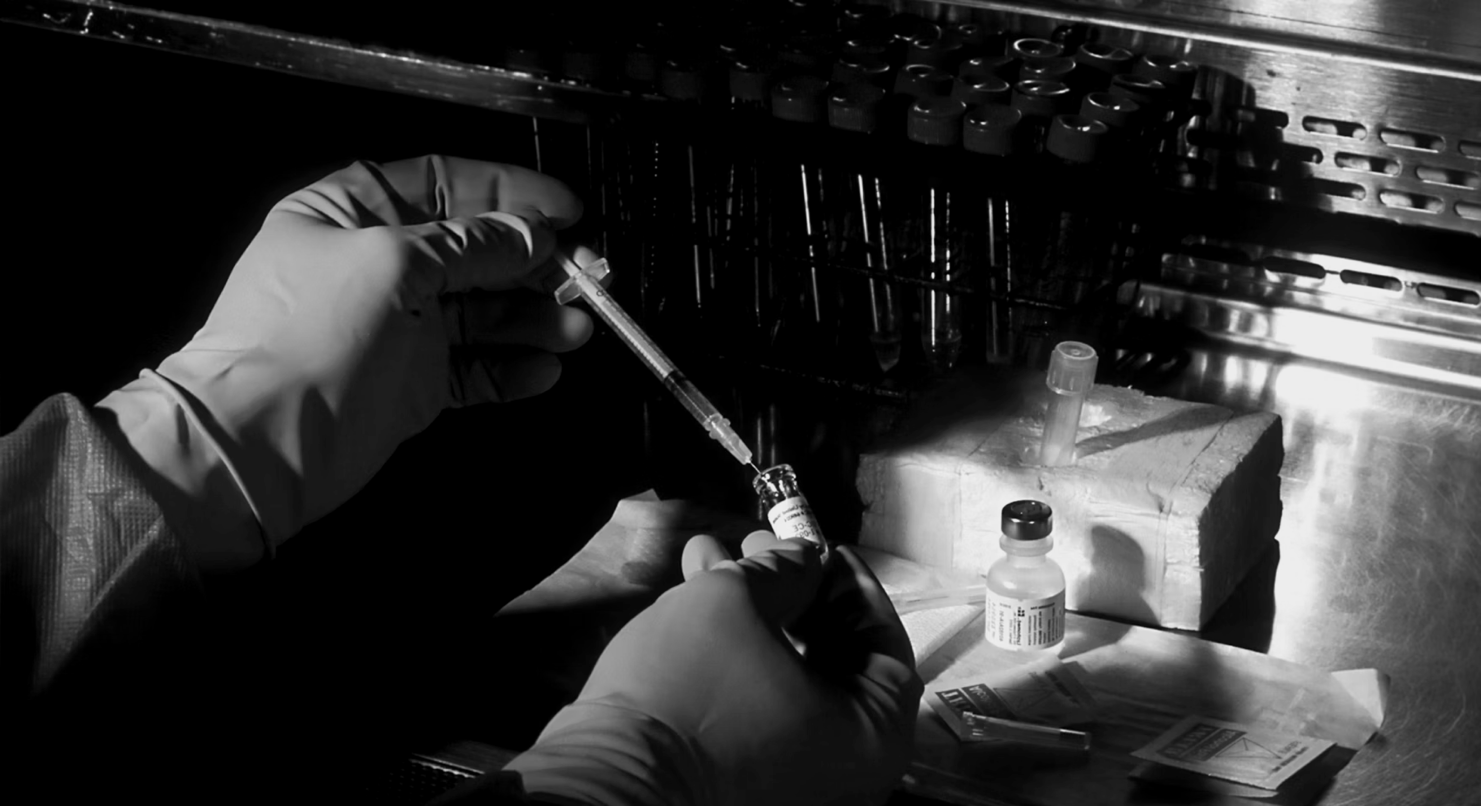 Gloved hands handling a syringe and vial in a lab setting with test tubes and medical supplies nearby.