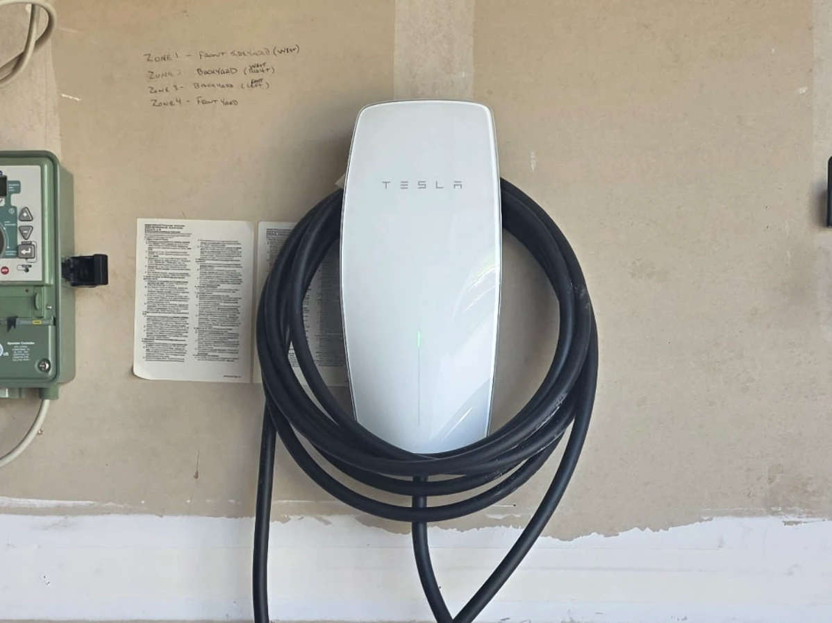 Tesla Wall Connector with its cable coiled, mounted on a garage wall beside a disconnect and subpanel