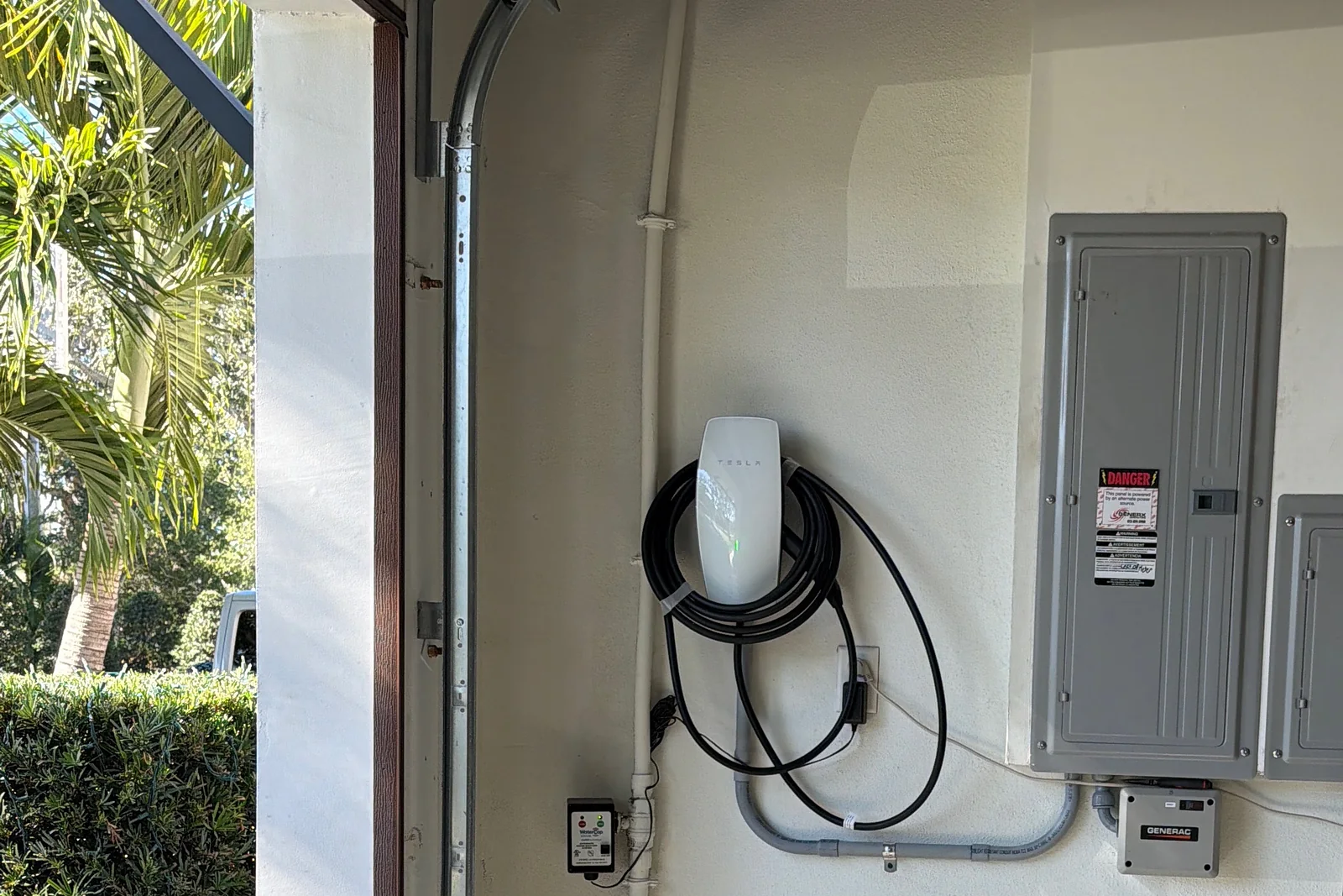 Tesla Wall Connector installed in a home garage next to the electrical panel, with palm trees outside the open door