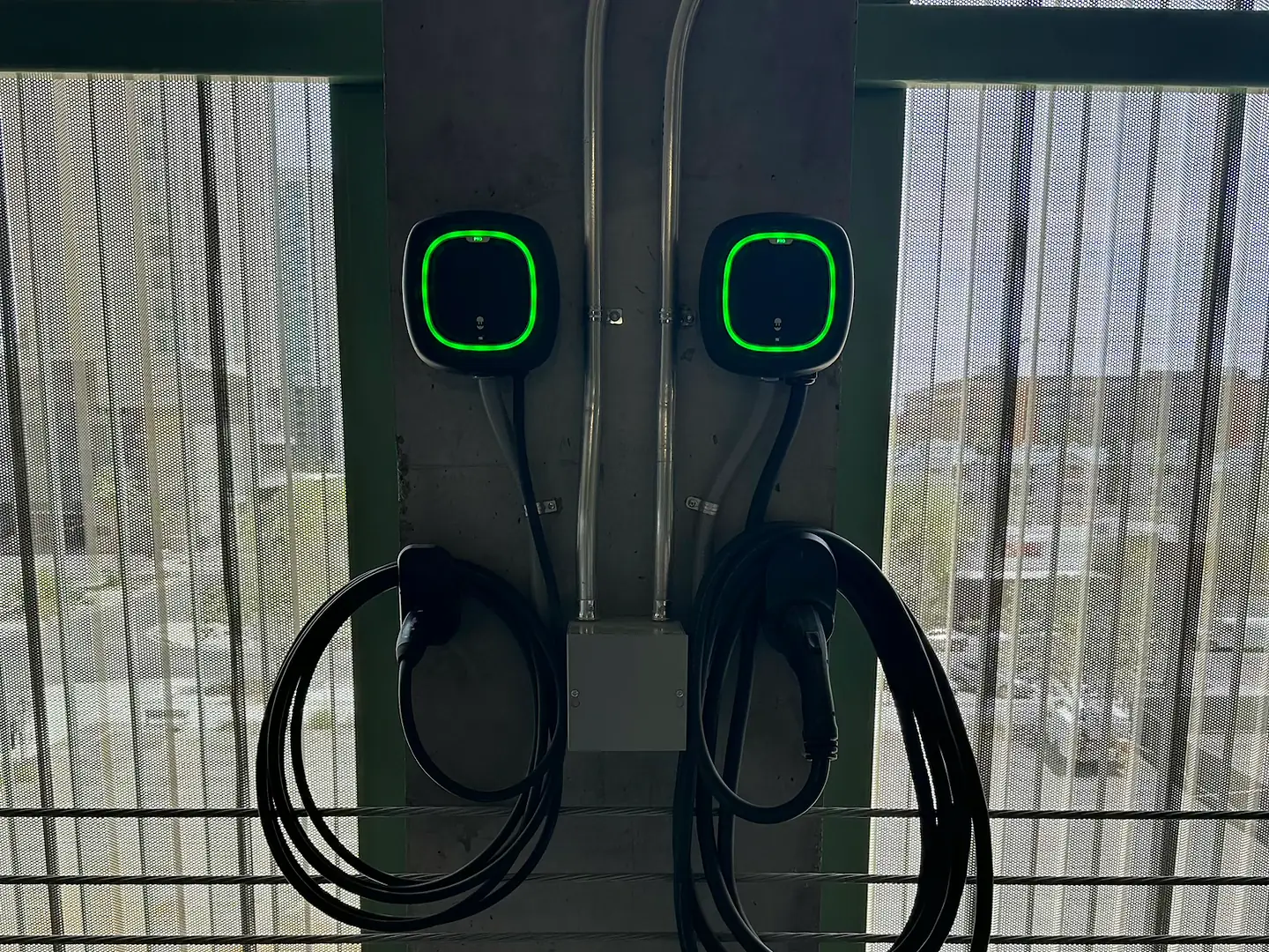Two wall-mounted EV chargers with green status rings on a parking structure column, fed by conduit from above