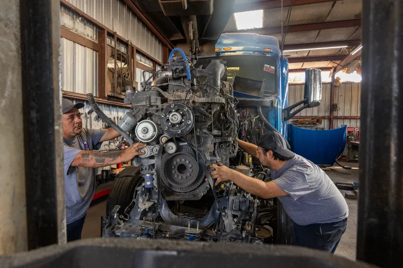Two mechanics perform engine replacement on a semi-truck, guiding a suspended engine into position.