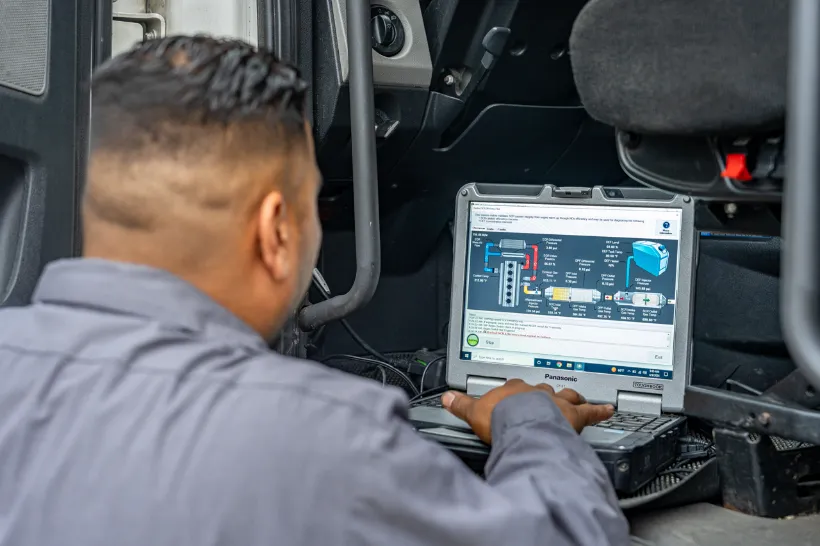Technician performs electrical system maintenance using diagnostic software on a rugged Panasonic laptop inside a truck cab.