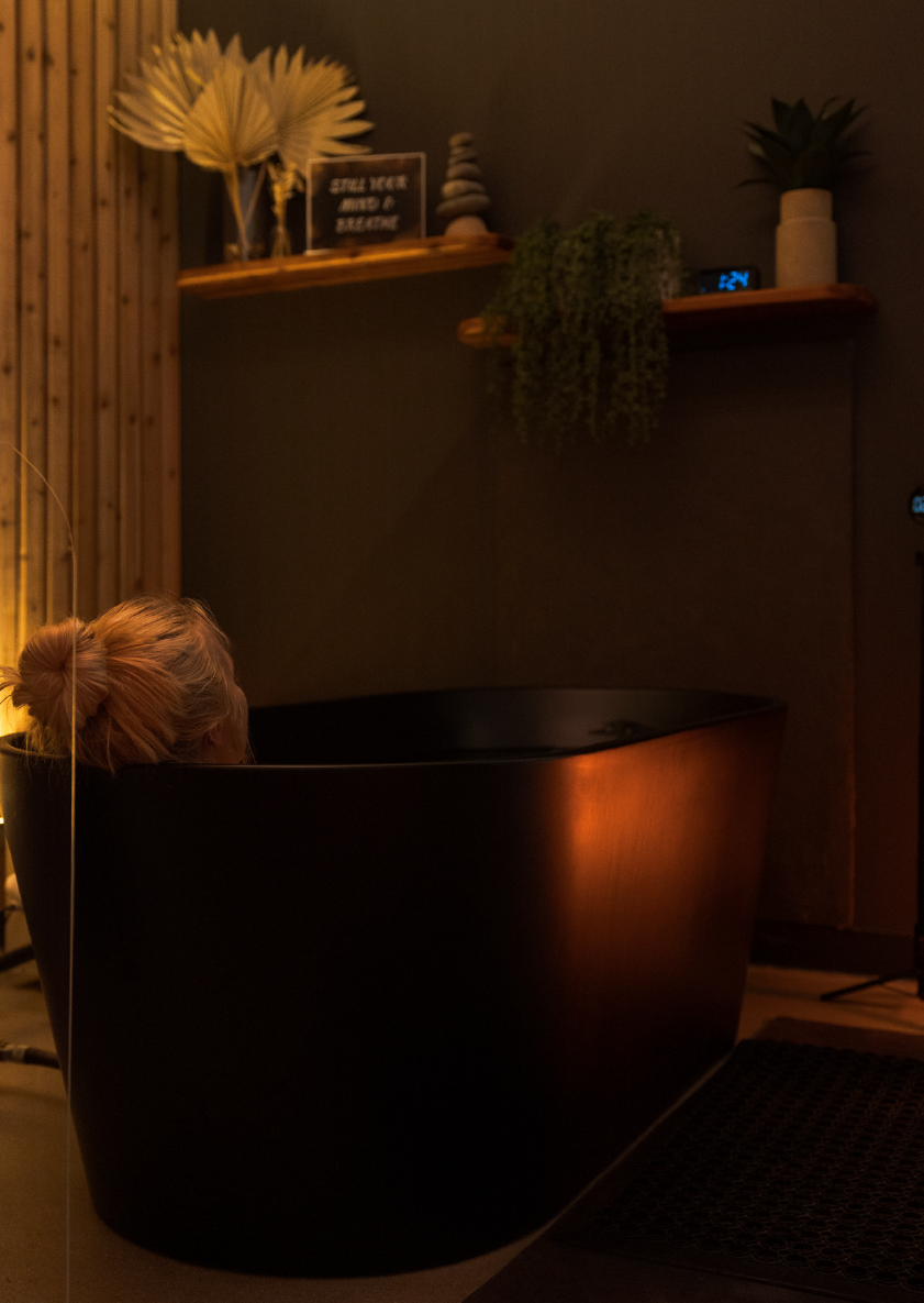Person with blonde hair relaxing in a dark freestanding bathtub in a dimly lit room.