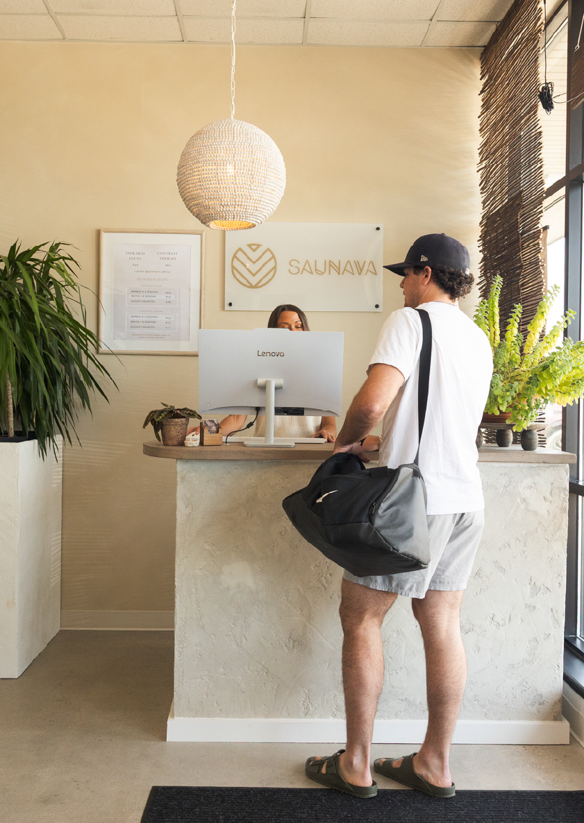 Man in casual clothes with gym bag at reception desk inside Saunava with plants and round light.