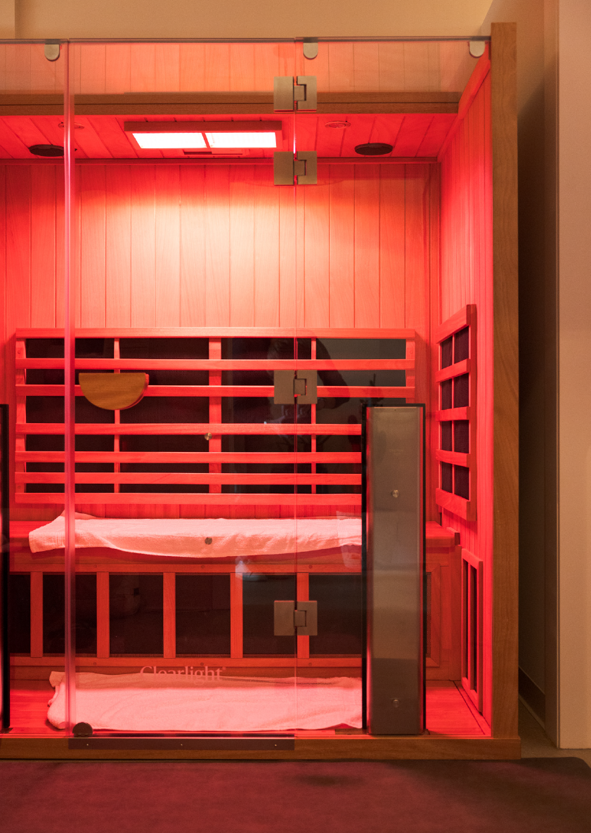 Wooden sauna room with glass door, two benches with towels, lit by red light.