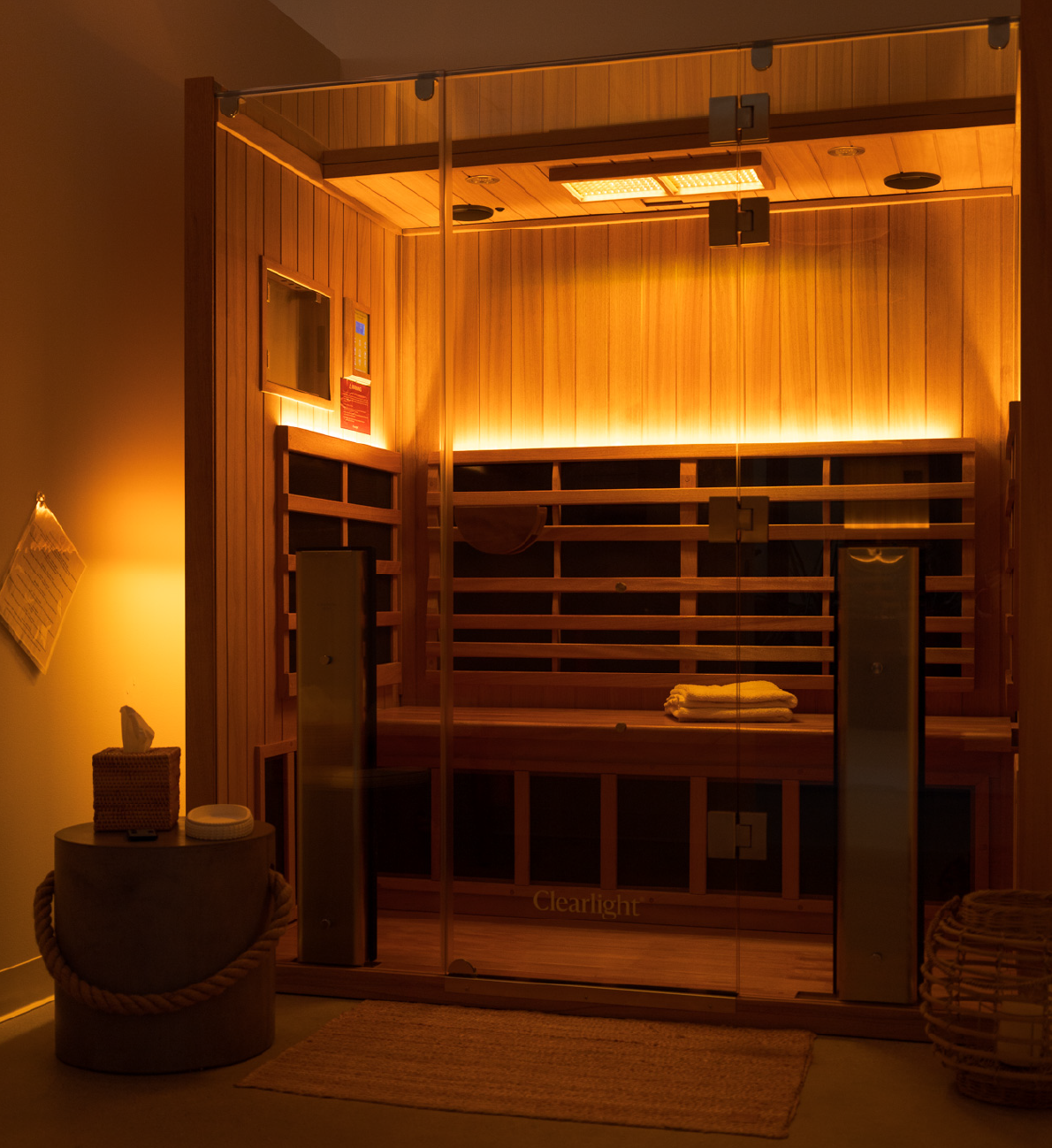 Warmly lit wooden sauna room with glass door and folded towels on the bench inside.
