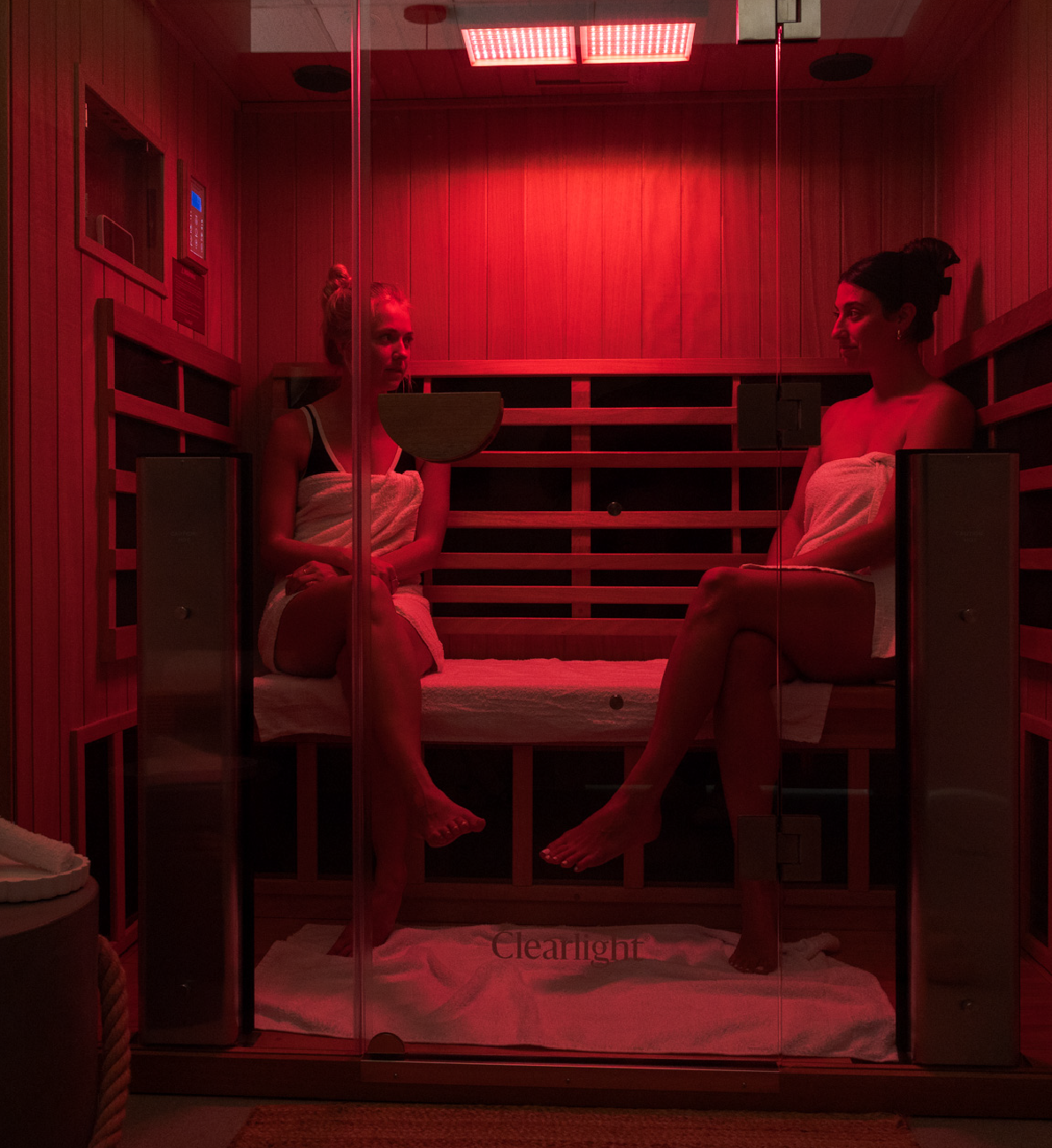Two women wrapped in towels sitting inside a red-lit wooden sauna room with glass door.