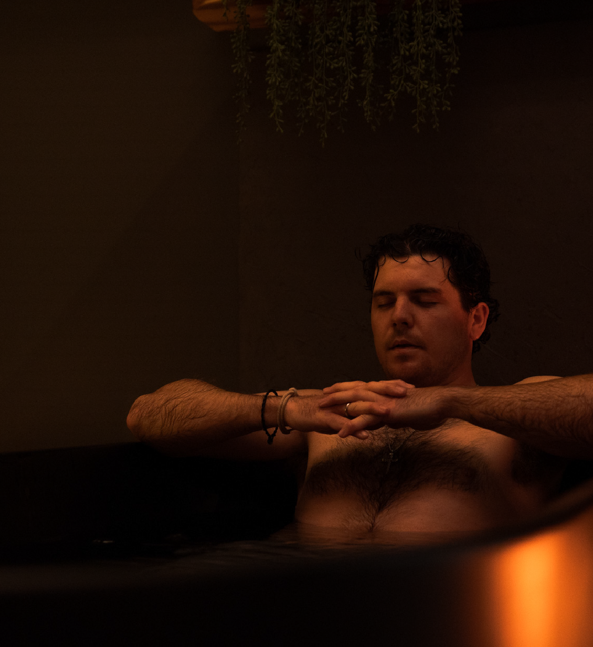 Man with closed eyes relaxing shirtless in dimly lit tub with hands clasped together.