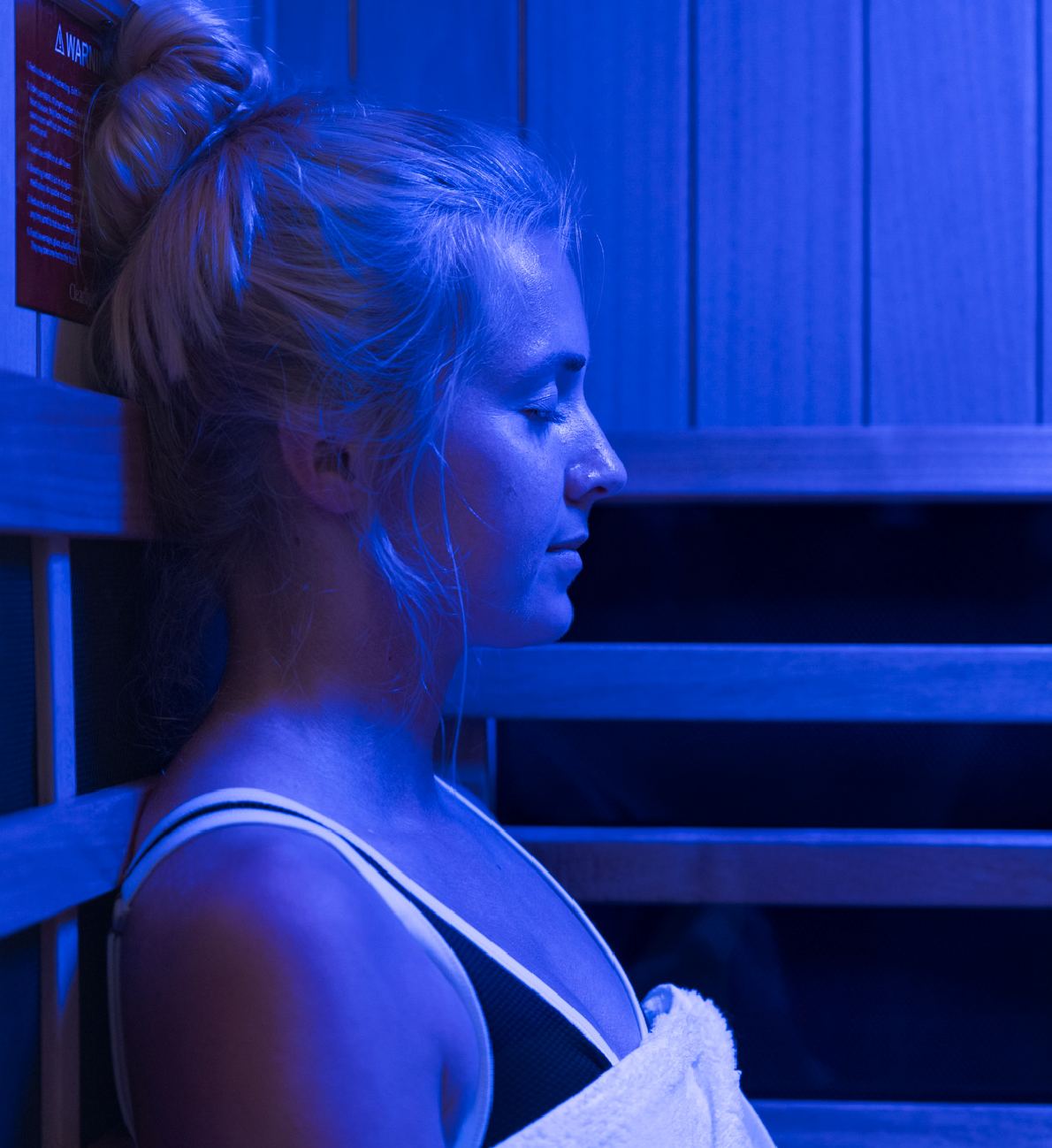 Woman with hair in bun relaxing with closed eyes inside a blue-lit sauna.
