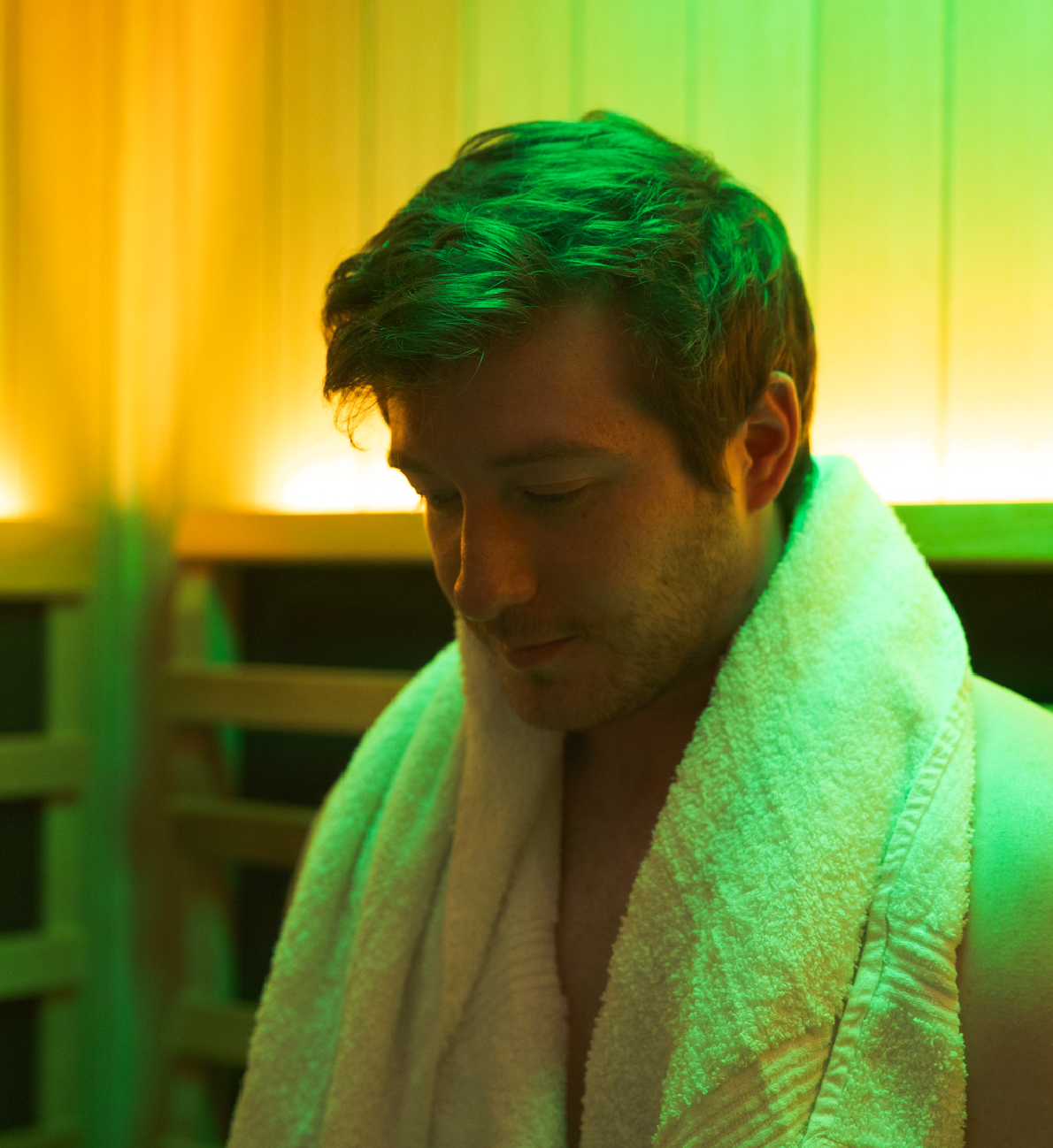 Man with a towel around his shoulders sitting in a sauna with warm backlighting.