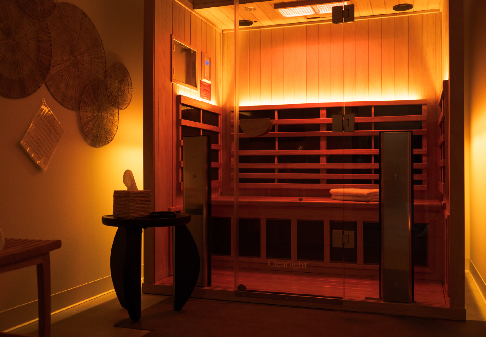 Wood-paneled infrared sauna with glass door, warm ambient lighting, and folded towels inside.