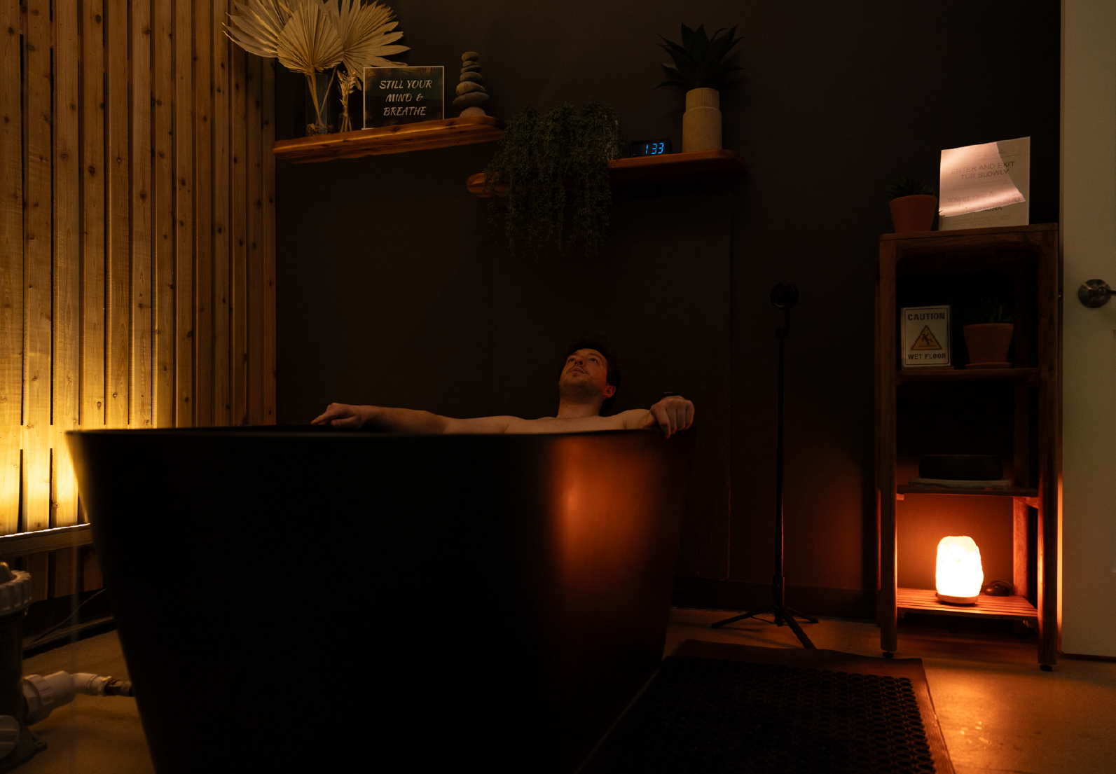 Man relaxing in a dark bathtub with ambient warm lighting and wooden decor in a spa-like room.