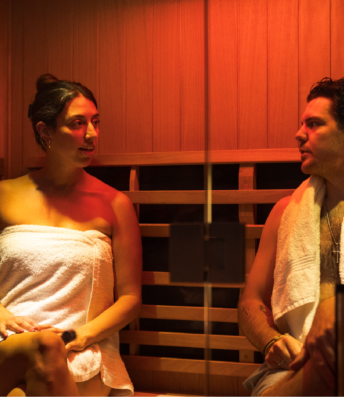 A man and woman sit wrapped in towels inside a wooden sauna, engaged in conversation.