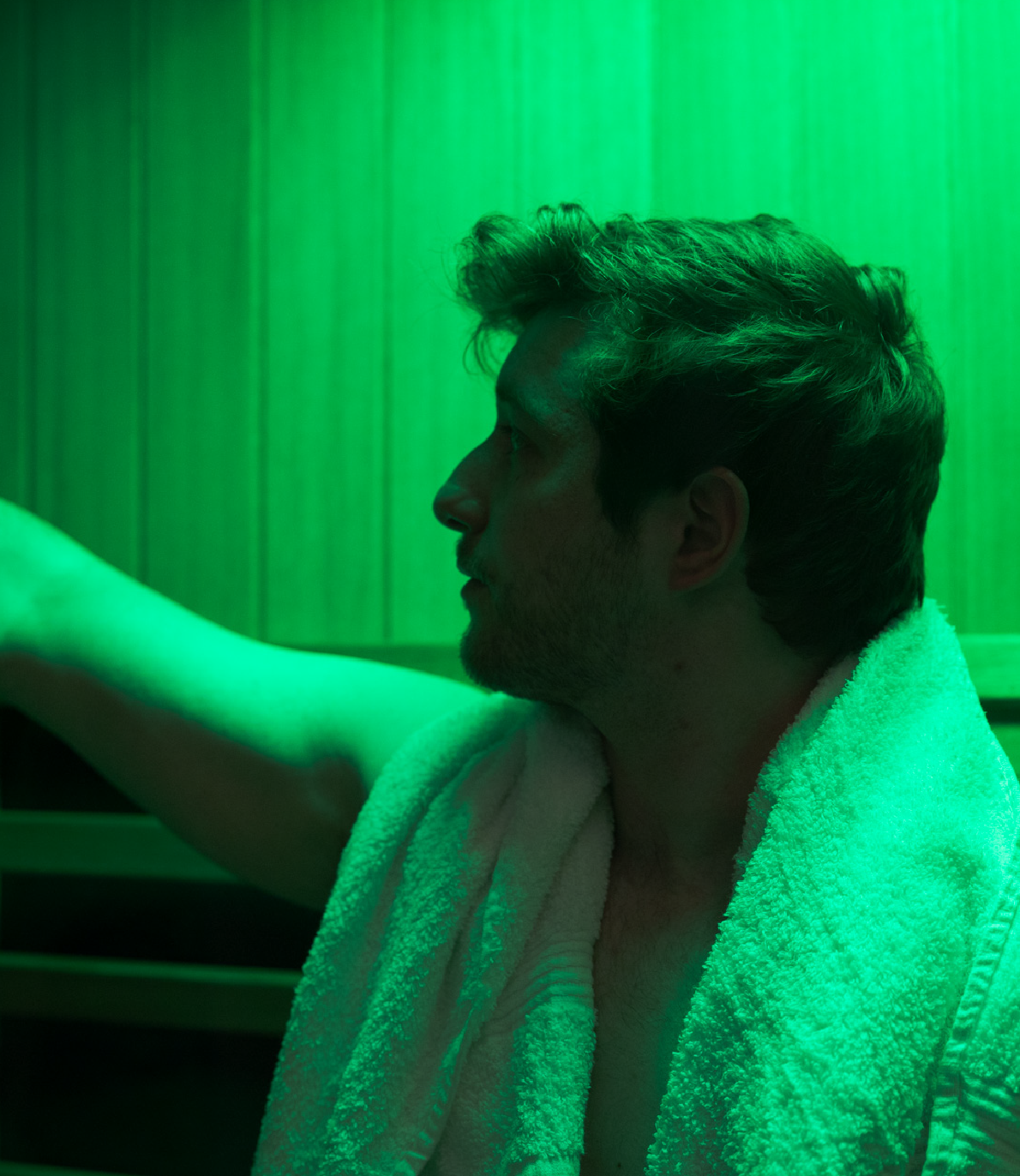 Man with towel around neck reaches out inside a green-lit wooden sauna room.