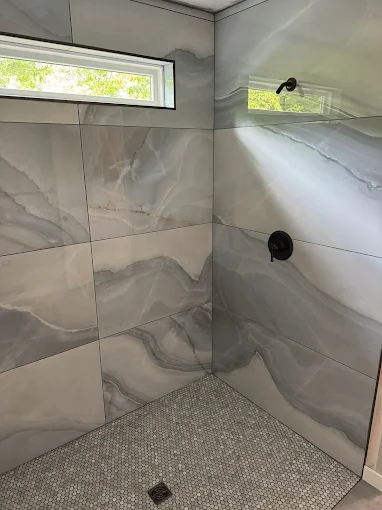 Corner shower with large gray marble-effect wall tiles, small hexagonal floor tiles, and a rectangular window.