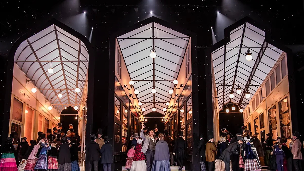 The glass-roofed Parisian arcade of Cafe Momus in La boheme at the Royal Opera House, crowd beneath falling snow.