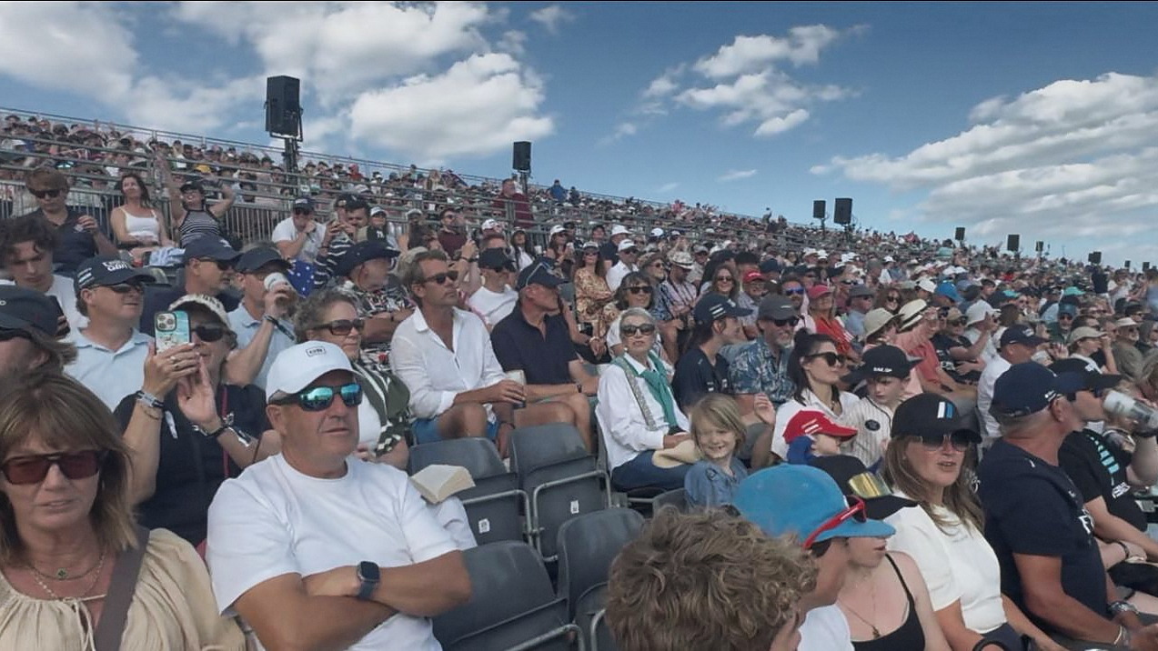 A packed temporary grandstand of spectators during racing, many in caps and sunglasses, under a bright sky.