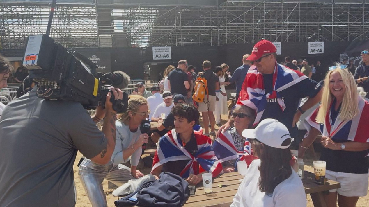 A broadcast camera operator and a presenter with a microphone record a group of supporters wrapped in Union flags at picnic tables behind the grandstand.