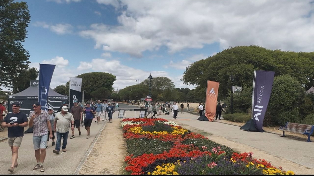 A wide path lined with municipal flower beds leads towards the race stadium, with sponsor flags and an official merchandise store on either side and visitors walking in both directions.