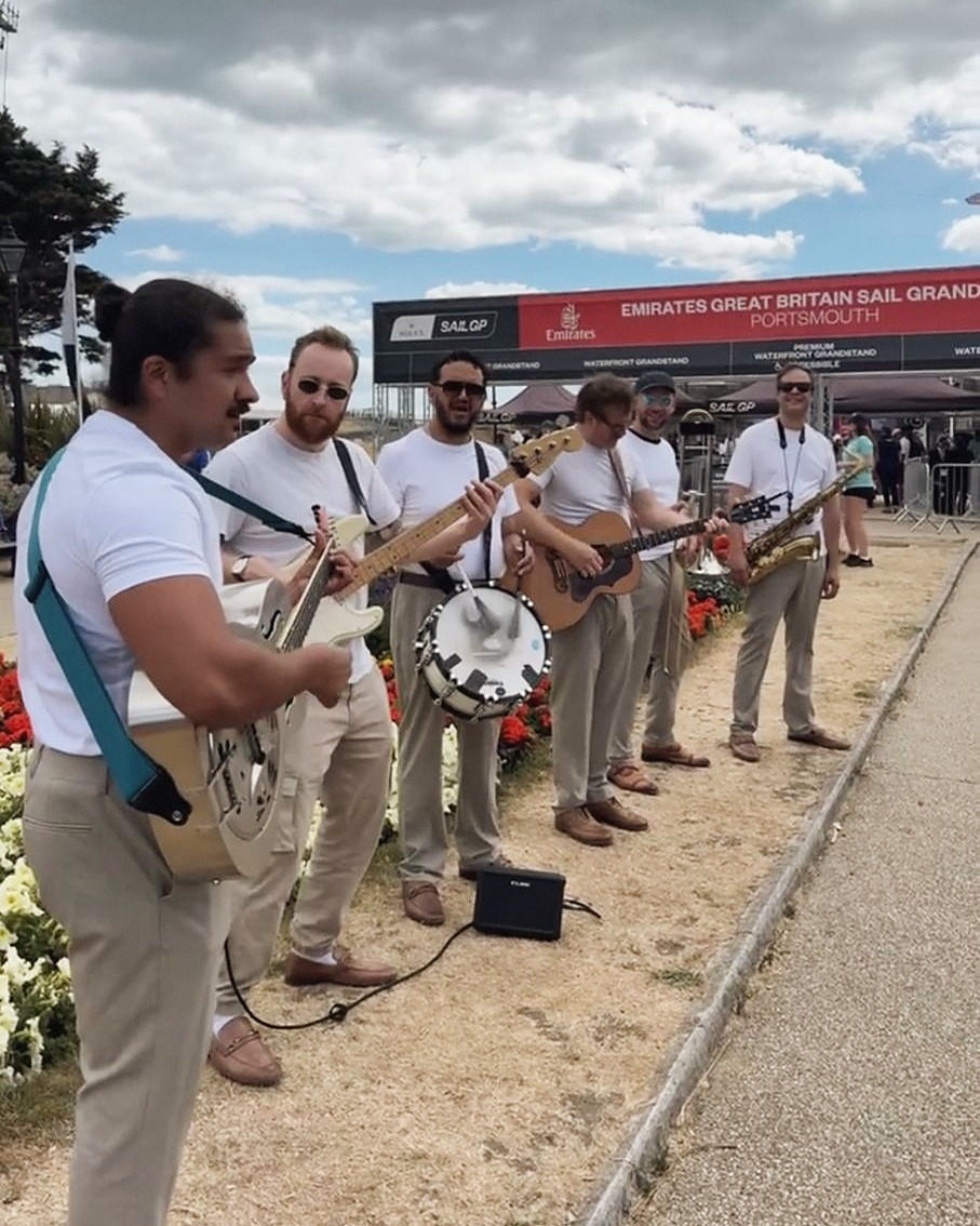 A band plays beside flower beds at the entrance to the race stadium, with tiered grandstand signage visible behind them.