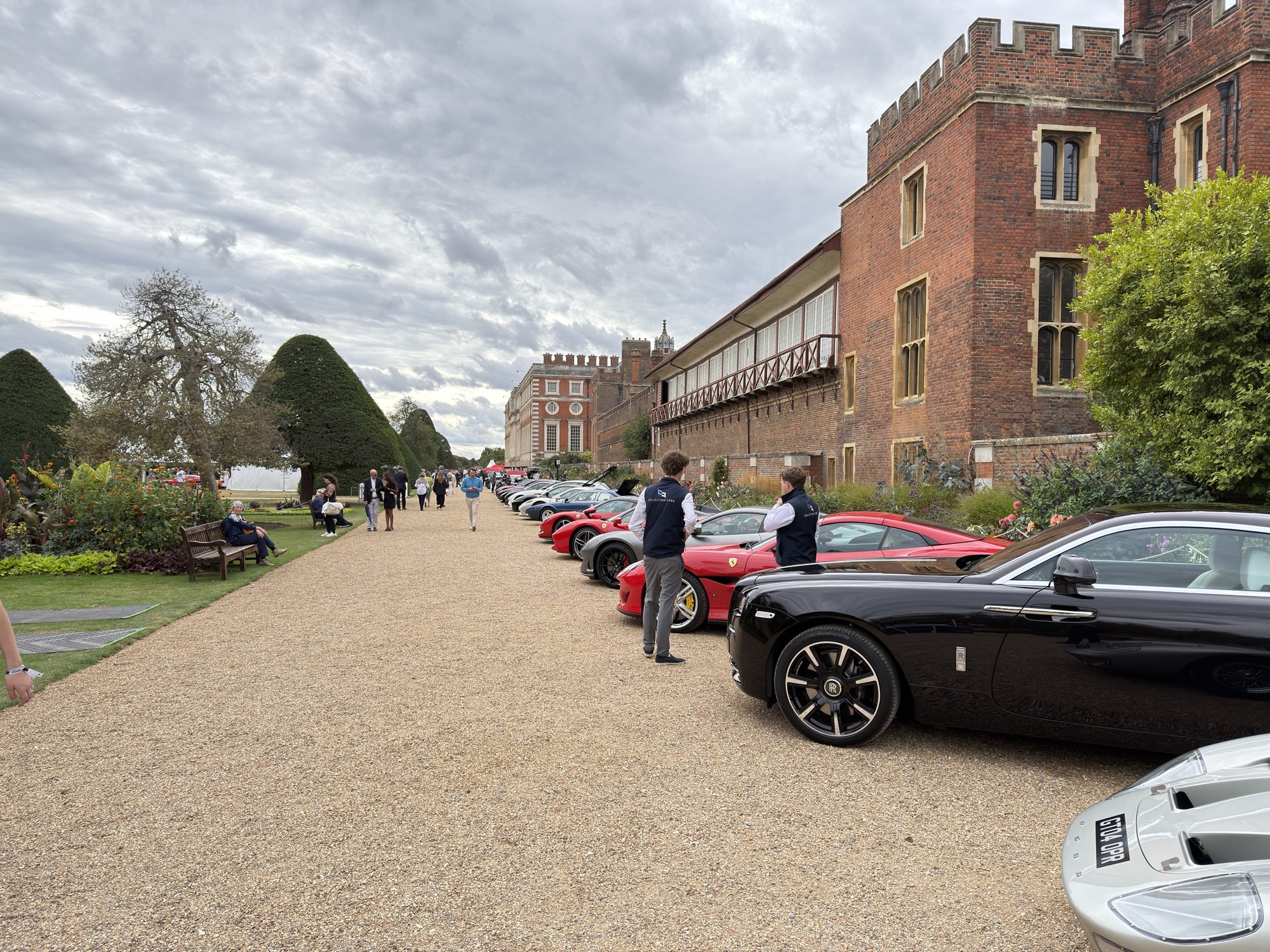 A line of collector cars — a black Rolls-Royce, Ferraris and a Ford GT — along a gravel path beside the brick wall of Hampton Court Palace under a grey sky.