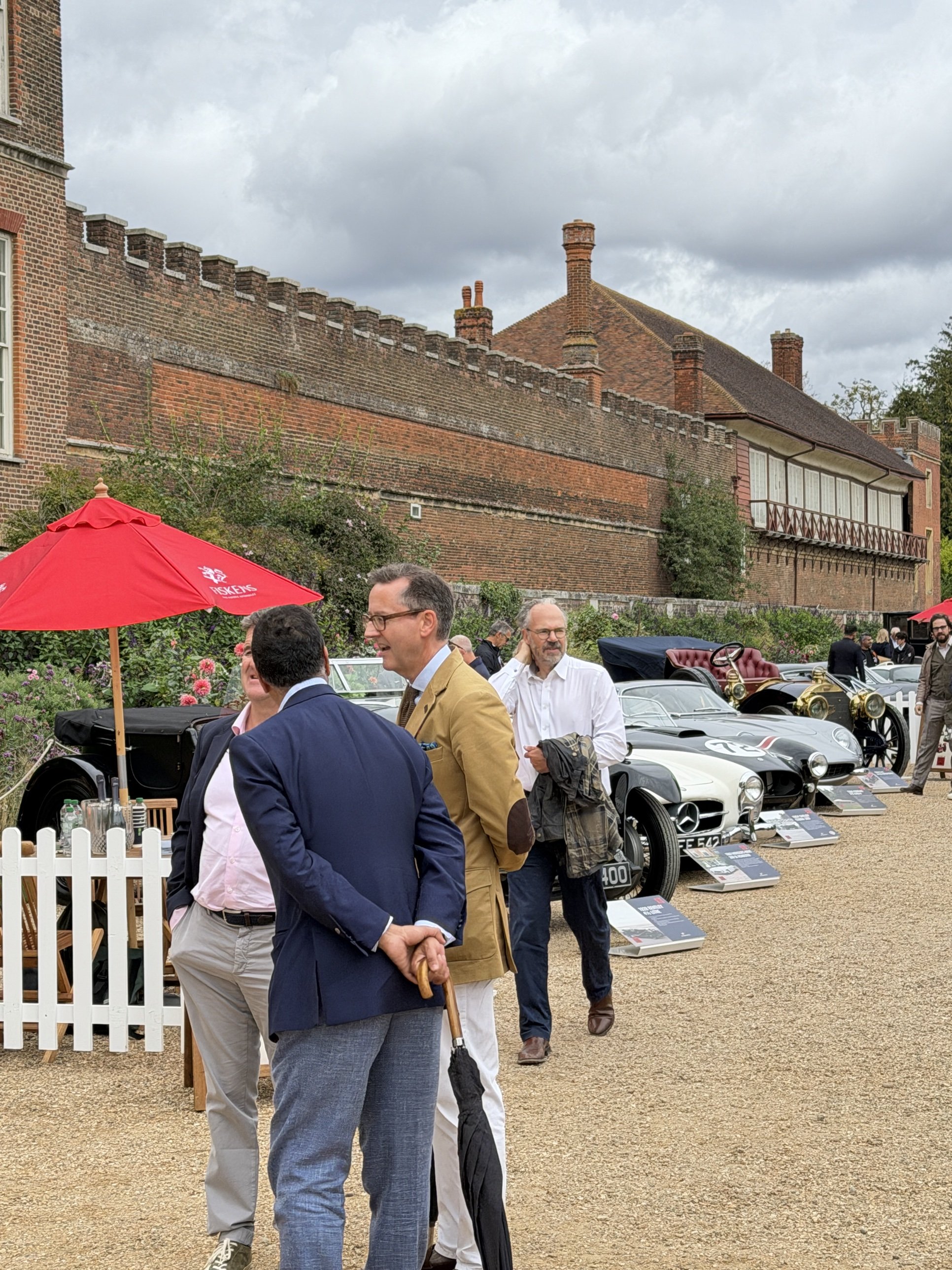 Guests in tailored jackets talking on the gravel beside a row of classic racing and veteran cars.