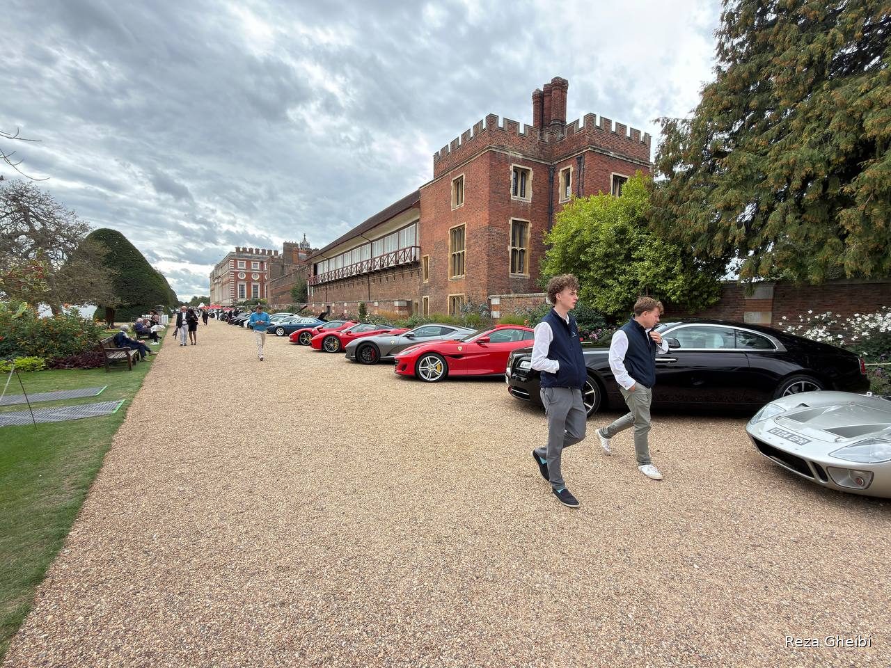A line of current supercars — Ferraris, a Rolls-Royce Wraith and a Ford GT40 — parked along the brick wall of Hampton Court Palace under a grey sky.