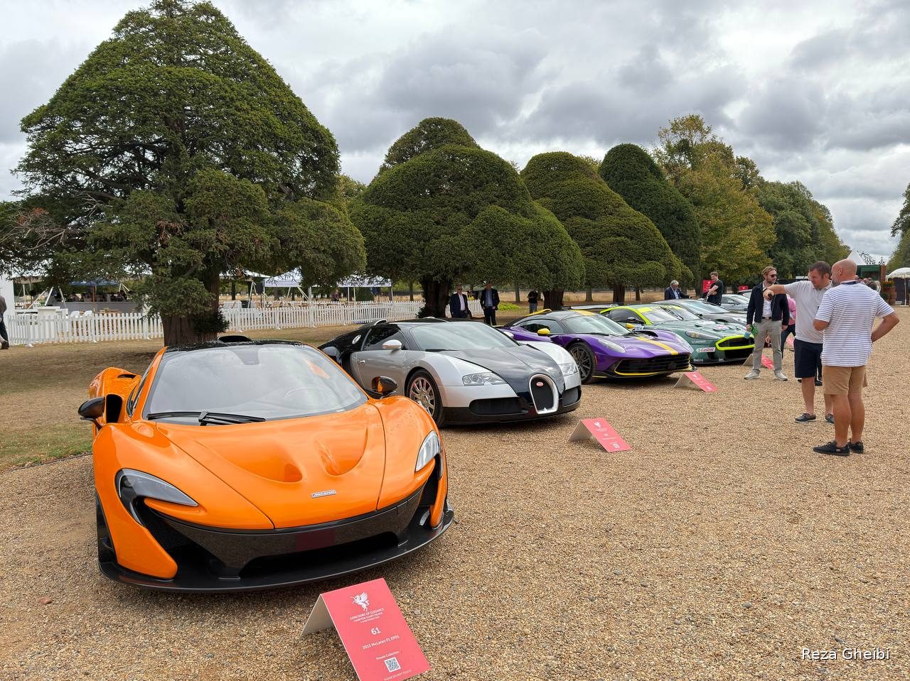 An orange McLaren P1 heading a row of hypercars parked under a clipped tree.
