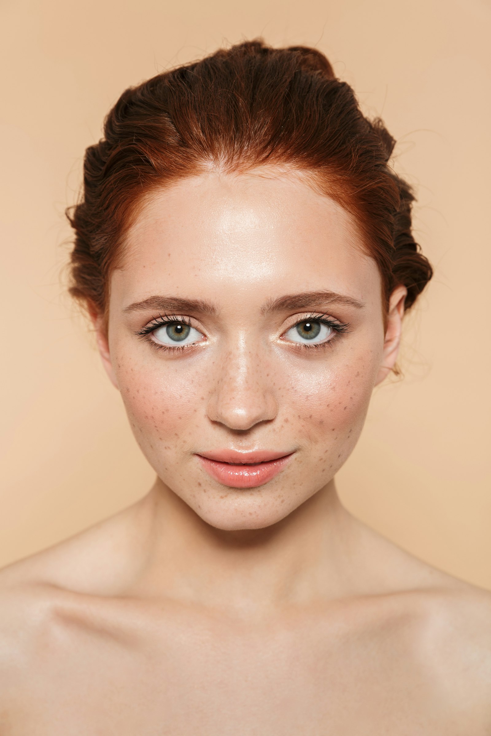 Close-up editorial portrait of a woman with auburn hair and freckles against a beige background.